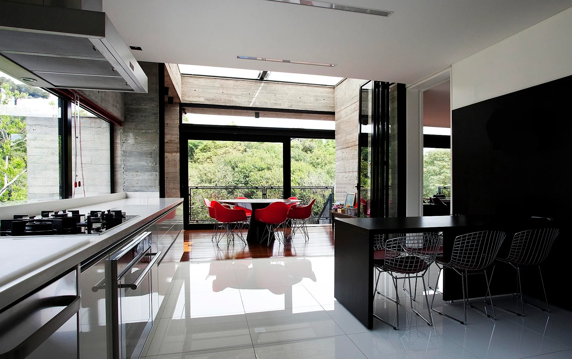 A modern open-plan kitchen features glossy white tiled floors, textured concrete walls, and a dining area with vibrant red chairs.