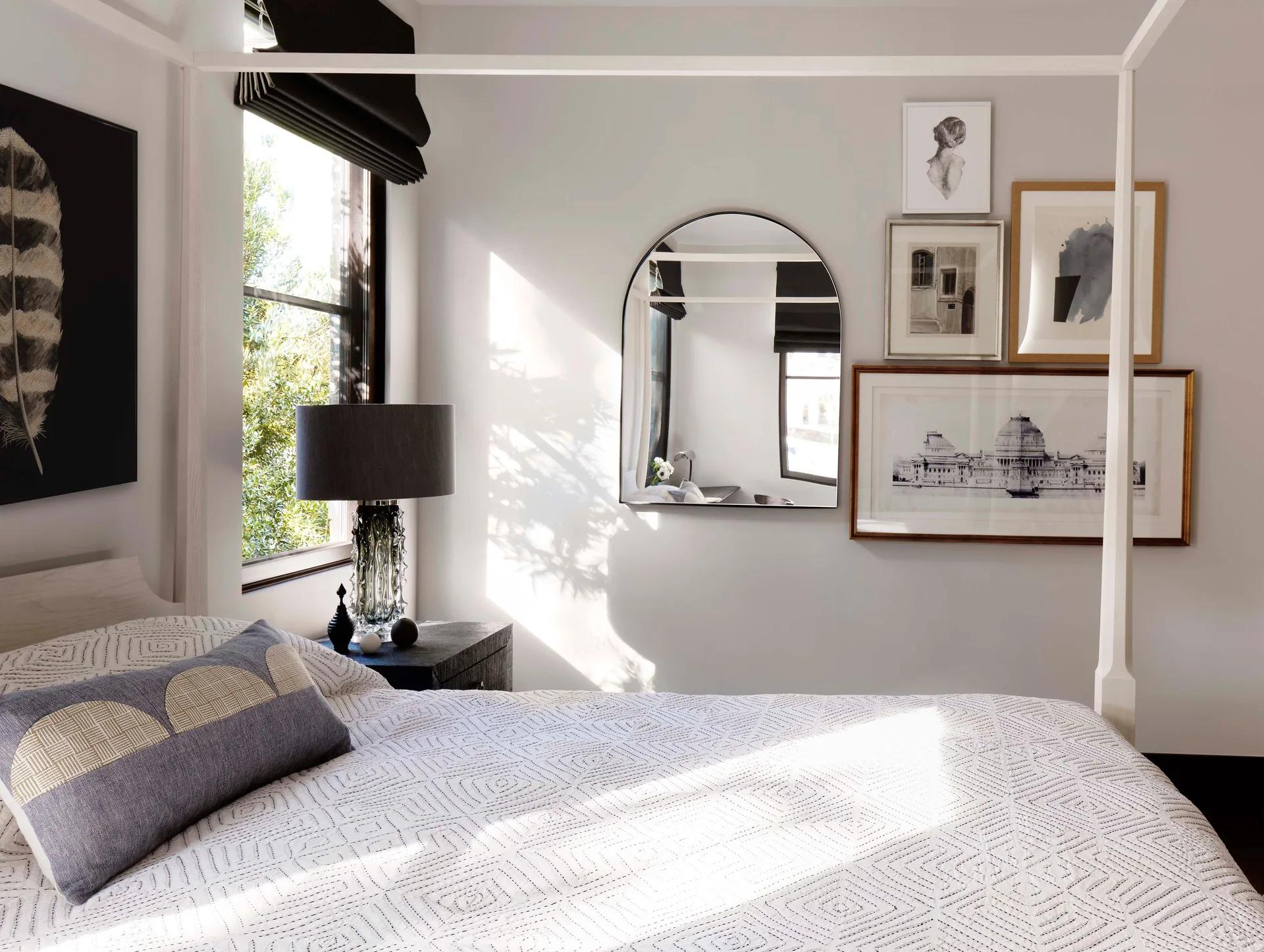 A bedroom with a white wooden canopy bed, a dark textured nightstand with a lamp, an arched wall mirror, and a small gallery wall of art.