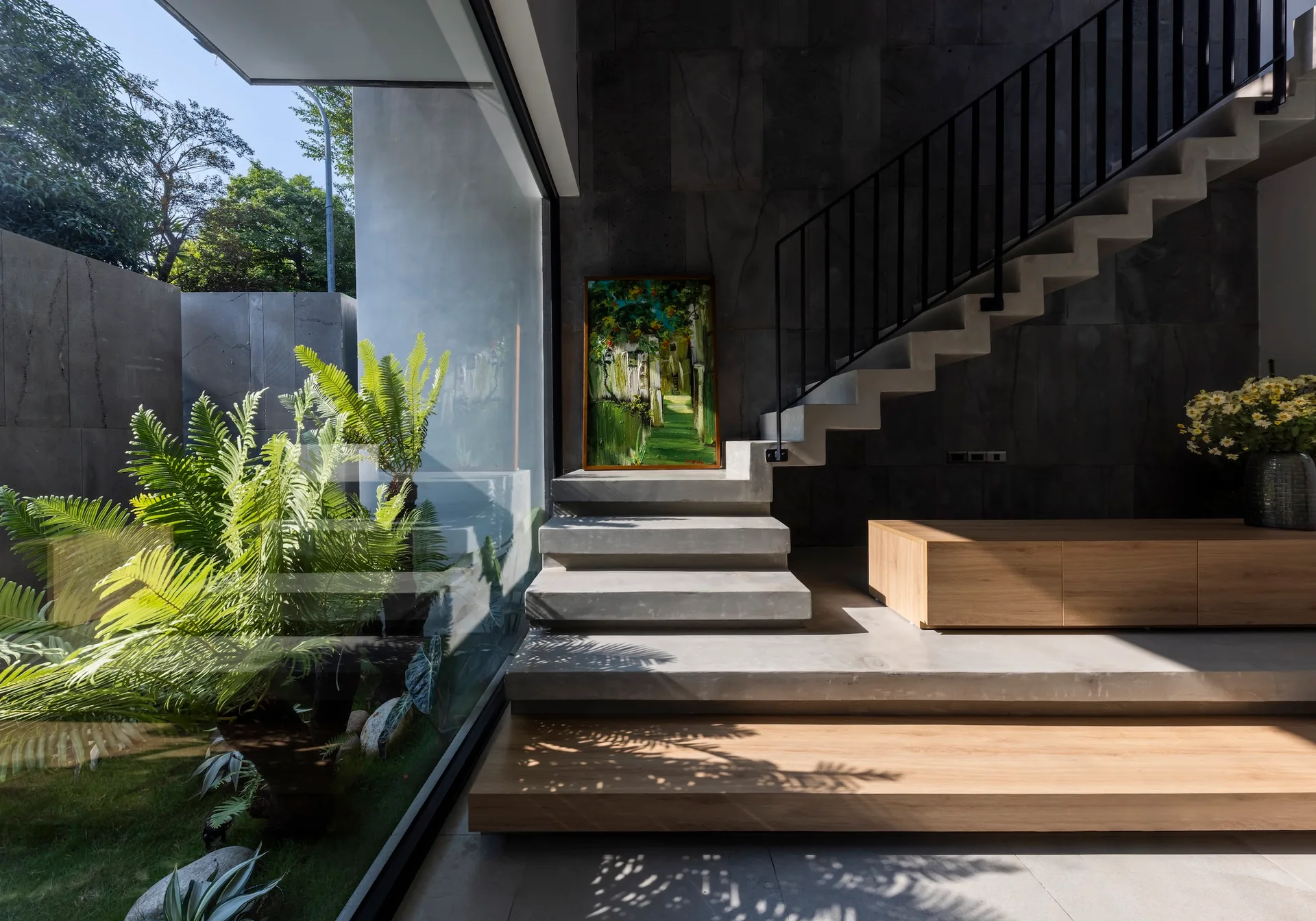 A minimalist concrete staircase with a black metal railing rises next to a large glass wall showcasing a lush green outdoor garden.