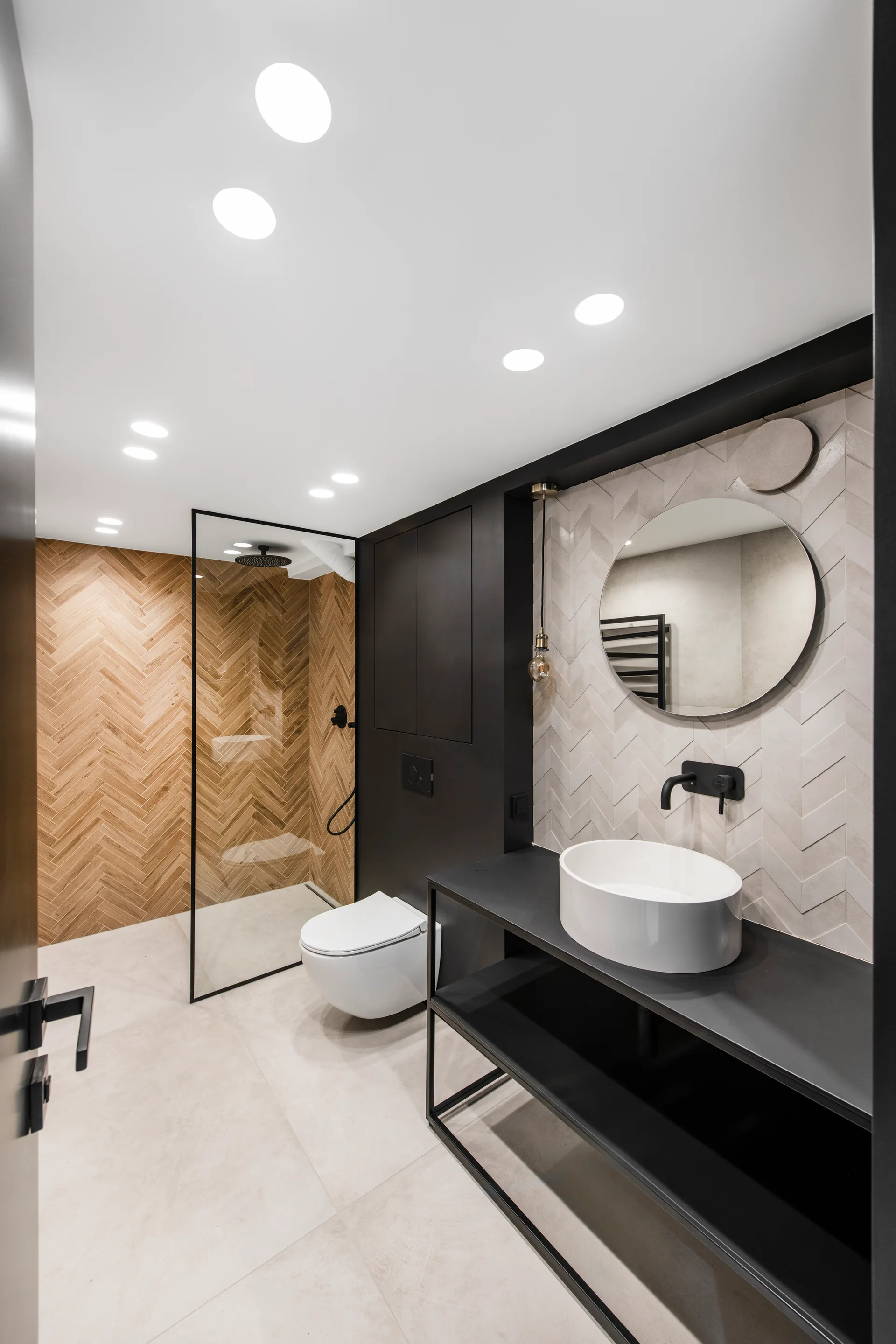 A modern bathroom featuring a black open-frame vanity with a white vessel sink, a round mirror, and walls tiled in a light herringbone pattern.