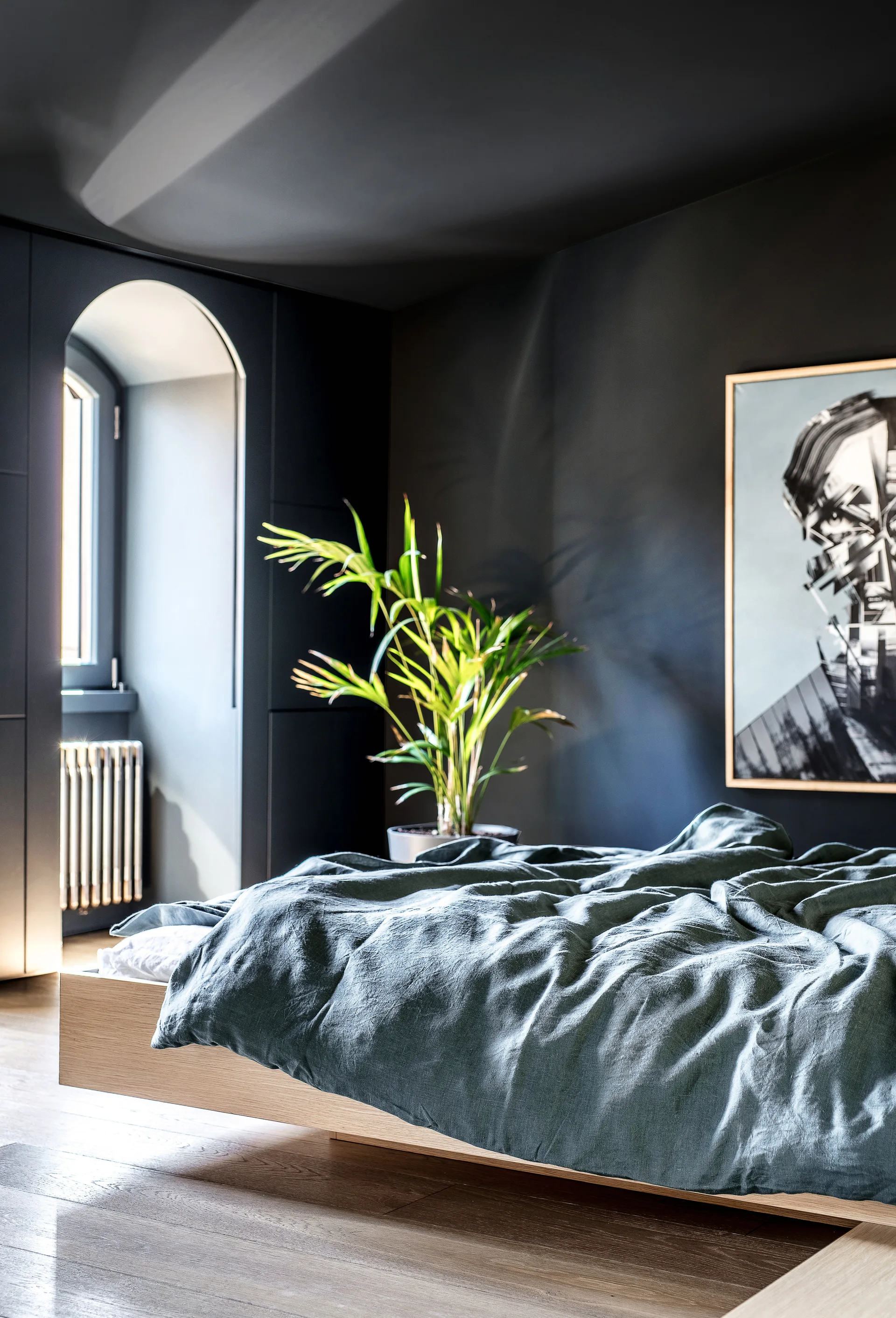 A modern bedroom with charcoal walls features a low wooden platform bed with blue linen sheets, a potted plant, and an arched window.
