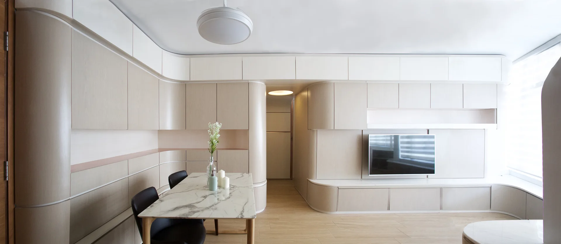 A minimalist living and dining room with seamless curved beige storage cabinets, a flat-screen television, and a marble-top dining table.