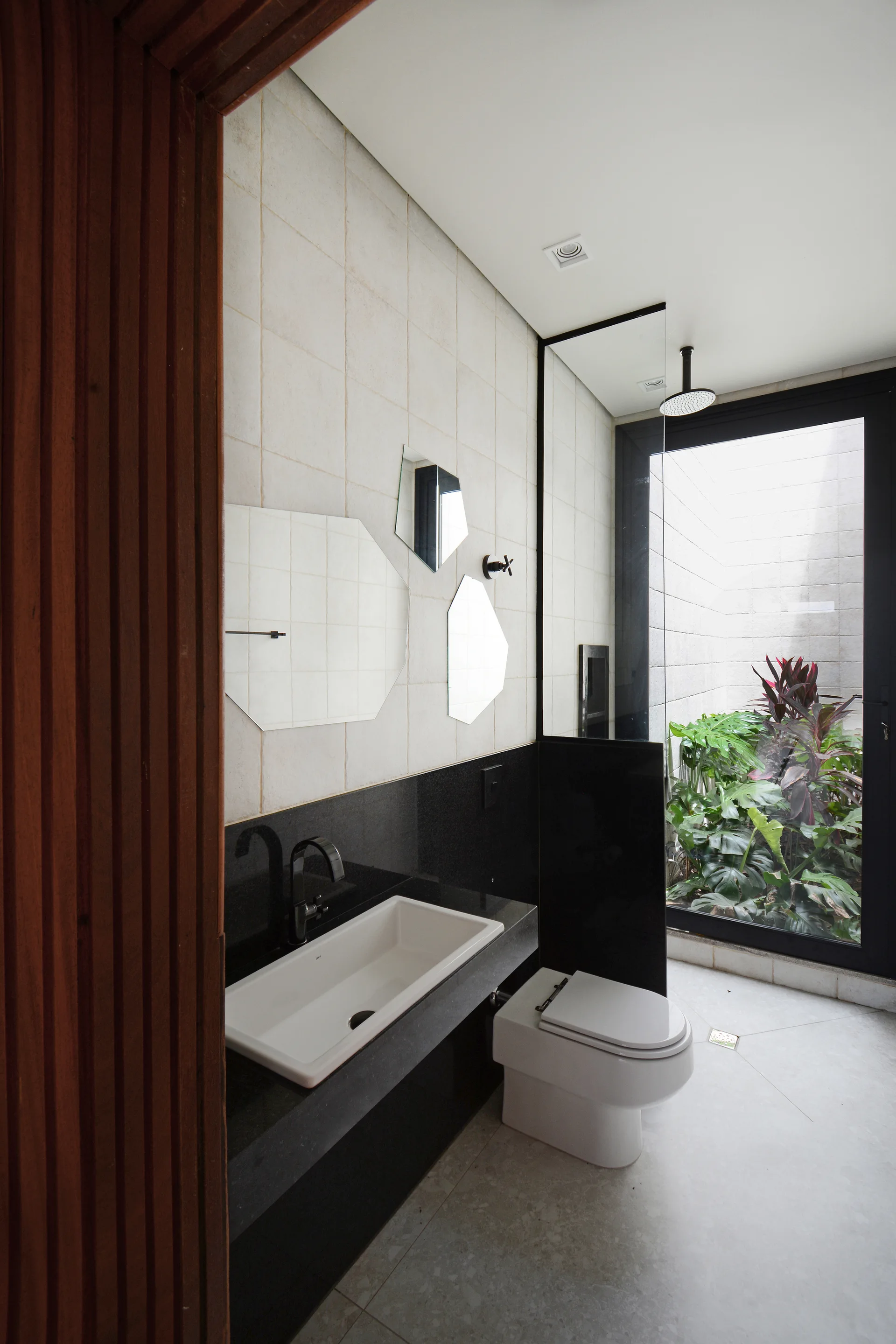 A modern bathroom featuring a black stone vanity with a white sink, geometric frameless mirrors, and a glass wall looking out to a lush green courtyard.