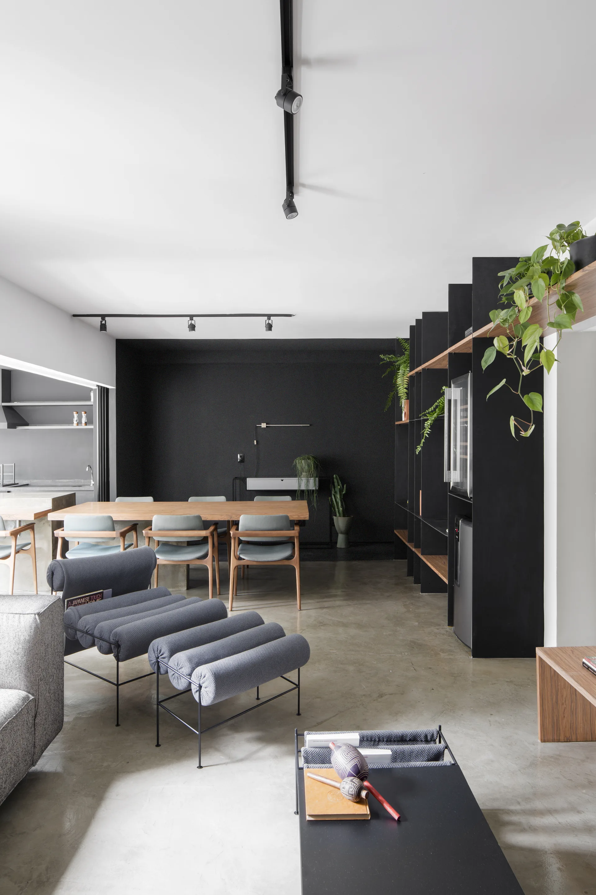 A contemporary open-plan living and dining room features a matte black accent wall, concrete floors, a wooden dining table, and a black shelving unit.
