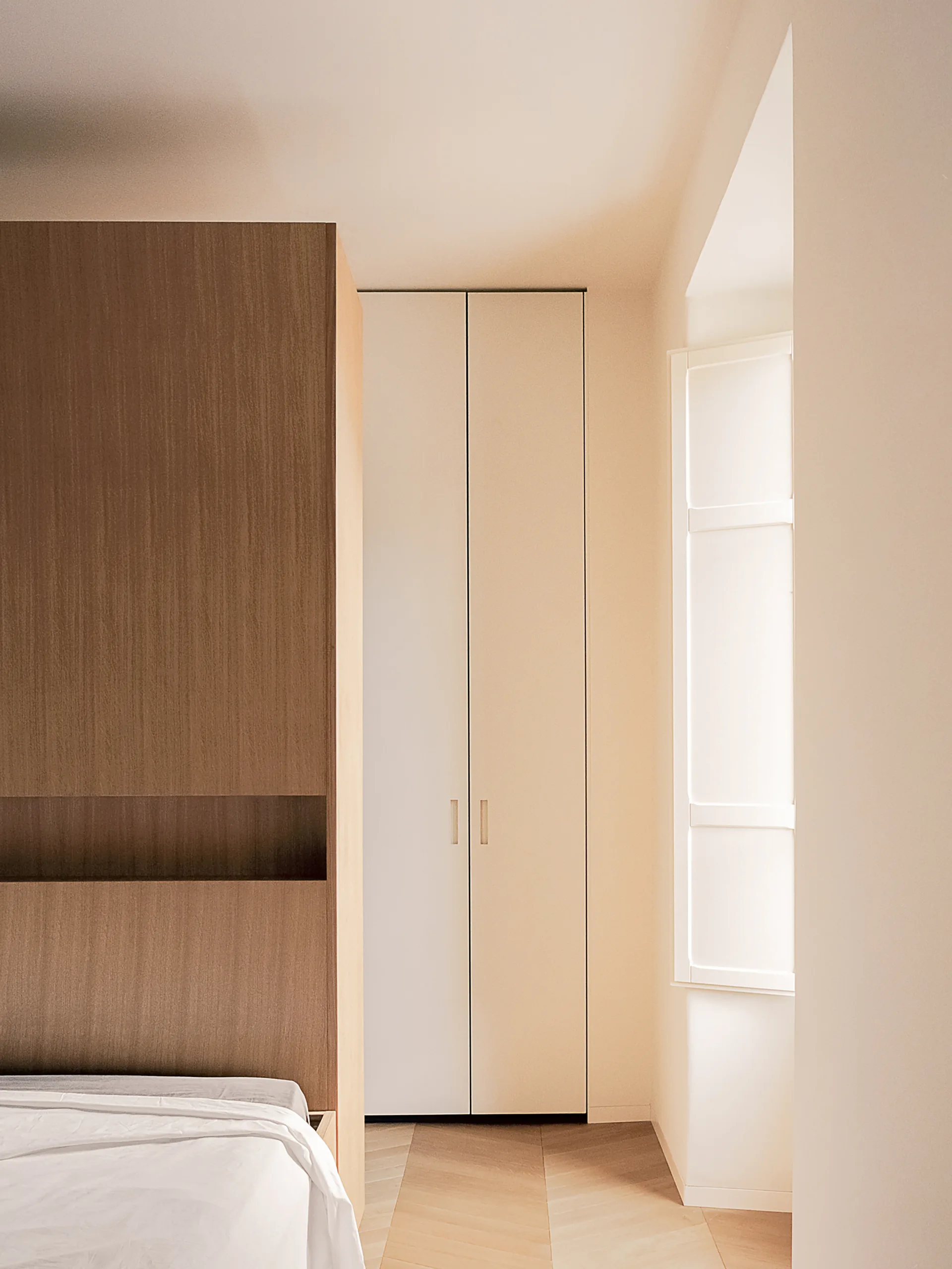 A minimalist bedroom featuring a tall white wardrobe, a wood-paneled headboard structure with a built-in niche, and light chevron pattern flooring.