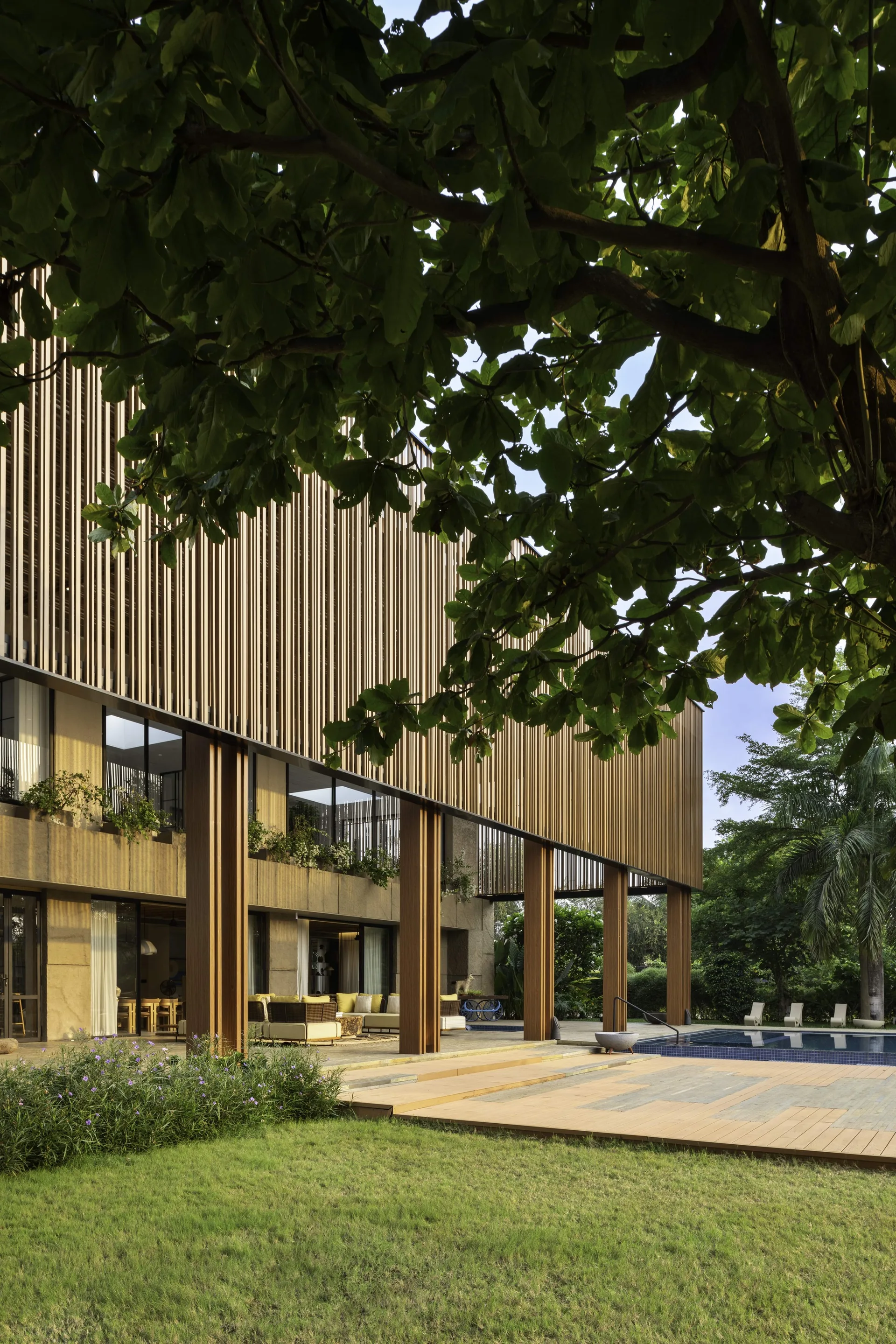 A contemporary rectangular swimming pool next to a wooden deck and a covered patio under a towering vertical wood slat facade.