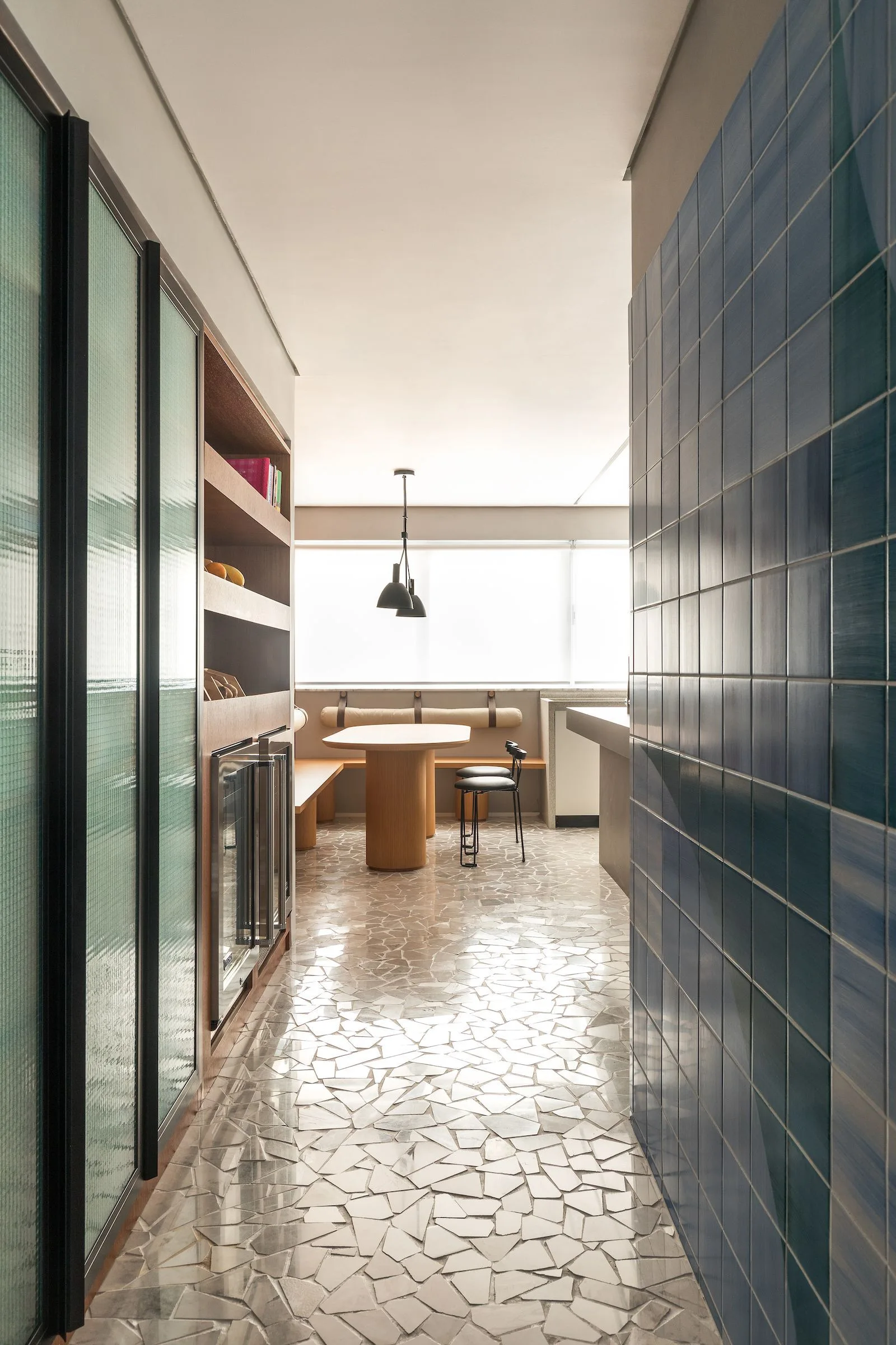 A long hallway with an irregular stone floor and a blue tiled wall leads to a modern dining area with a round wooden table and built-in bench.