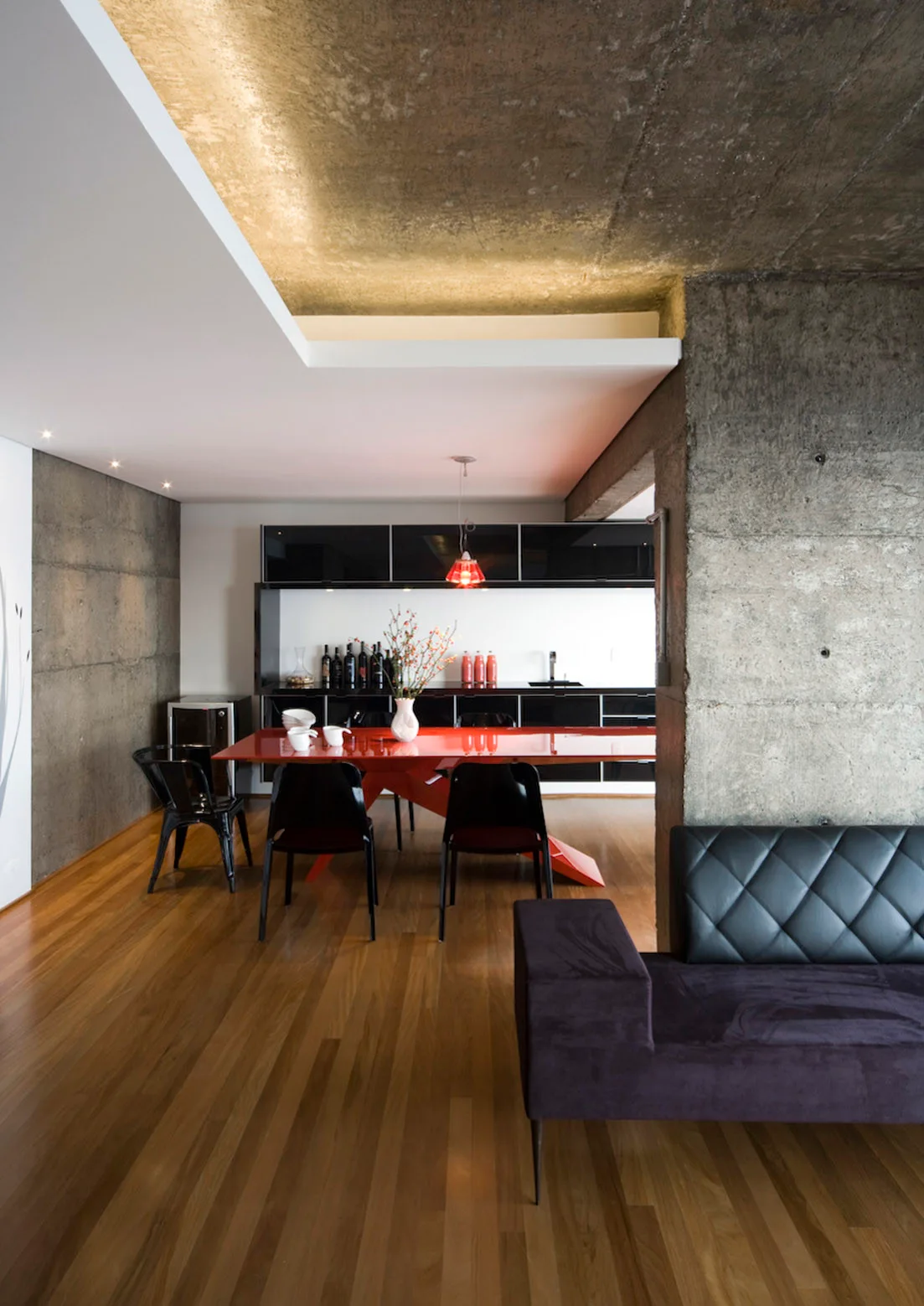 An open-concept living area features a bright red dining table, black chairs, glossy black cabinetry, and exposed raw concrete walls and ceiling.