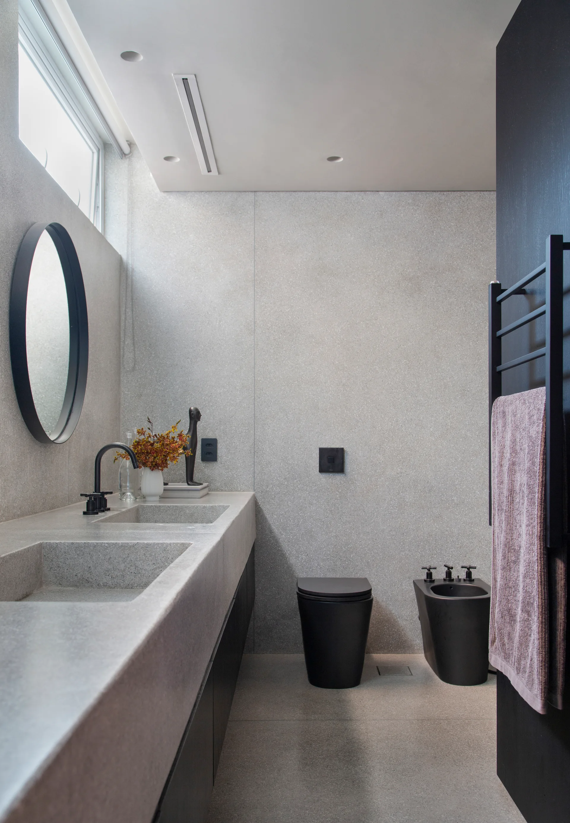 A modern minimalist bathroom with speckled grey stone walls, an integrated double vanity, a round mirror, and a matte black toilet and bidet.