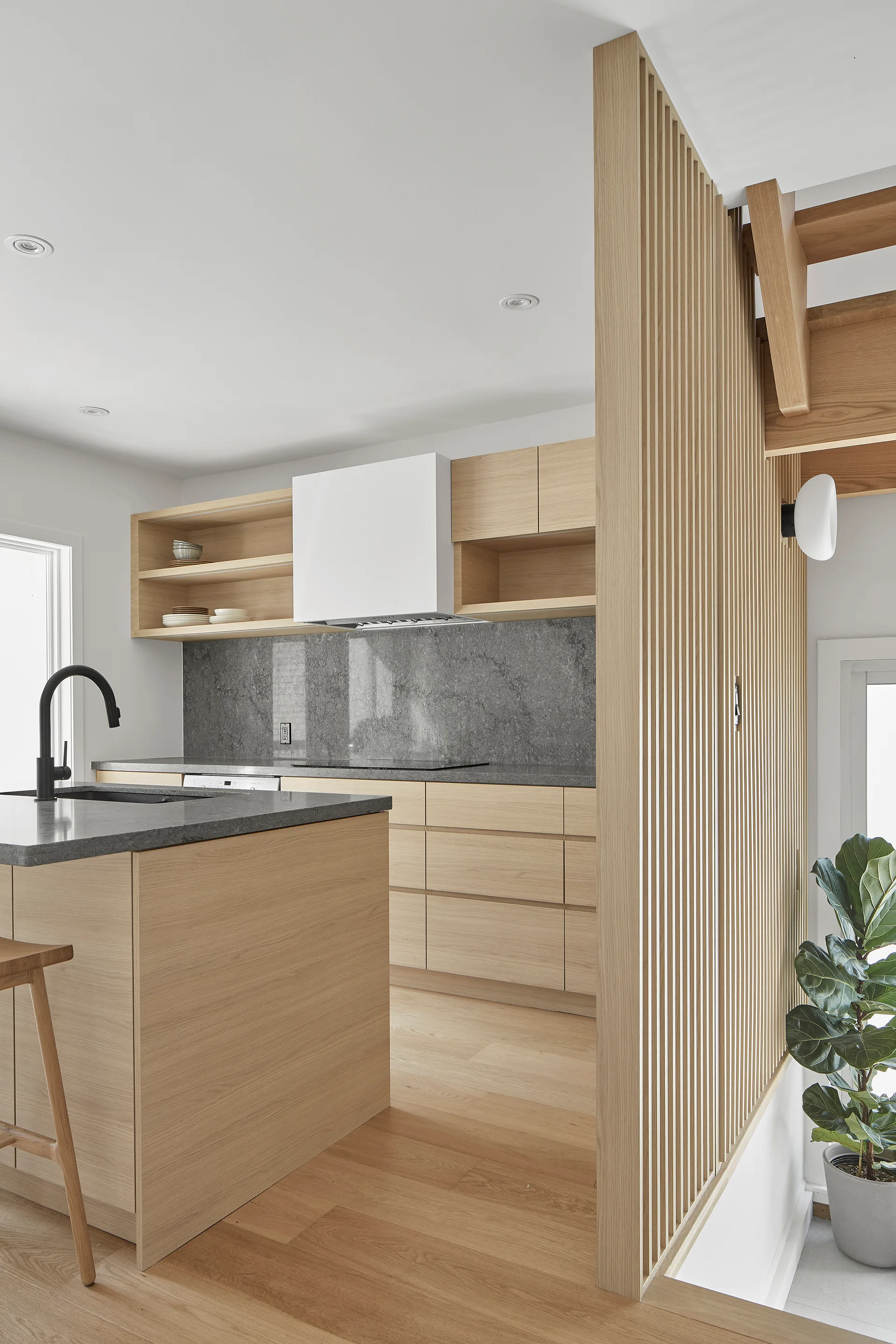 A modern kitchen features light wood cabinets, grey countertops, a black faucet, and a vertical slatted wood partition screening a staircase.