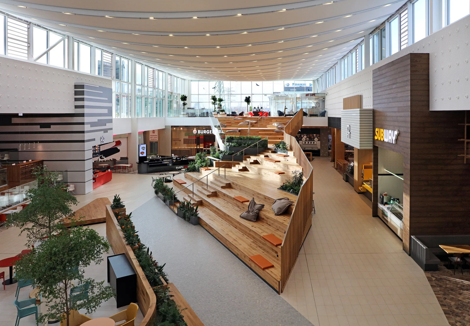 A spacious commercial atrium featuring a large wooden tiered seating structure with built-in planters, orange cushions, and surrounding storefronts.