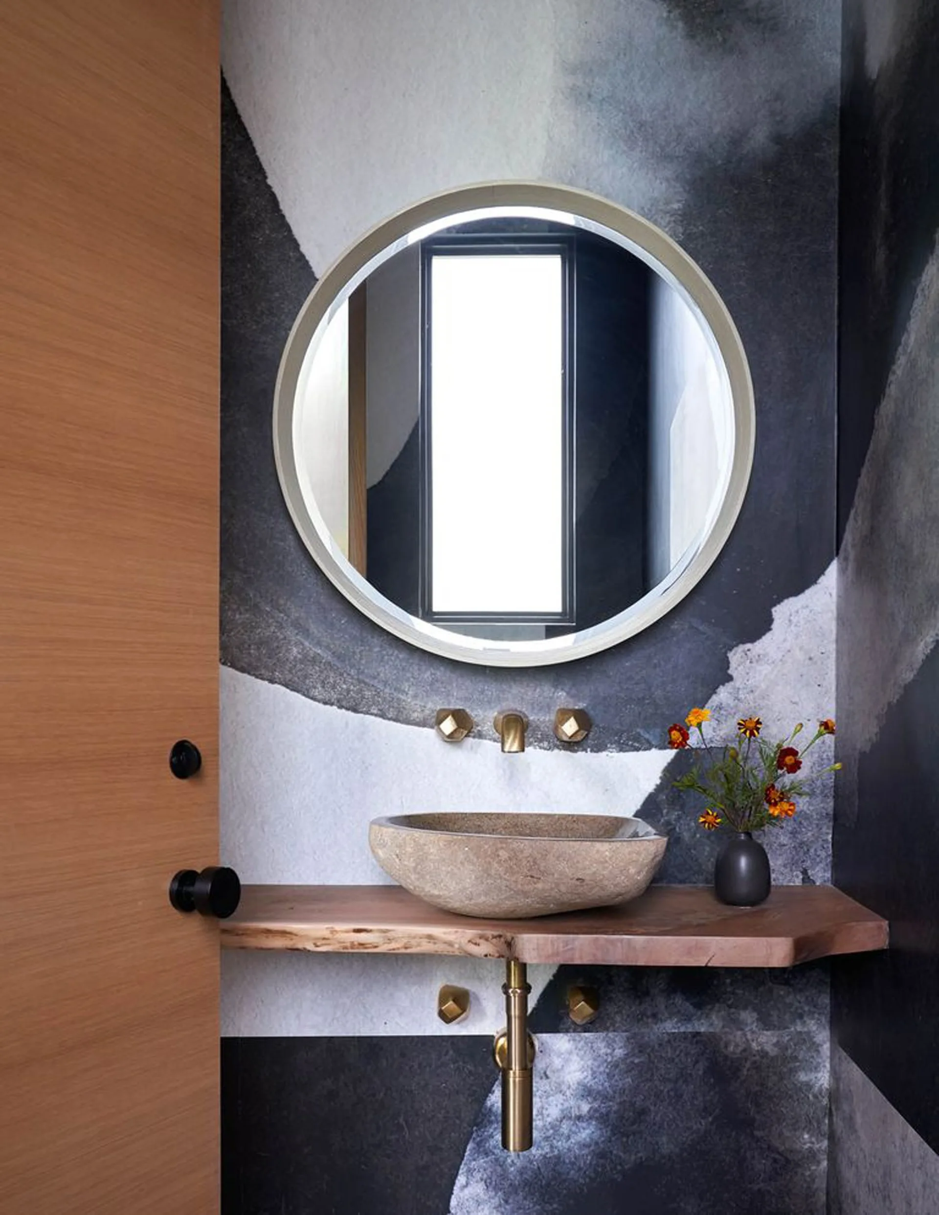 A modern powder room features a stone vessel sink on a live-edge wood vanity shelf against abstract black and white wallpaper.