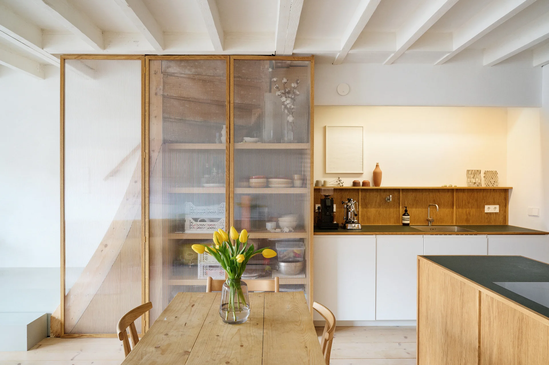 A minimalist kitchen and dining area features a wooden table with yellow tulips, white cabinetry, and a tall wood-framed cabinet with ribbed glass doors.
