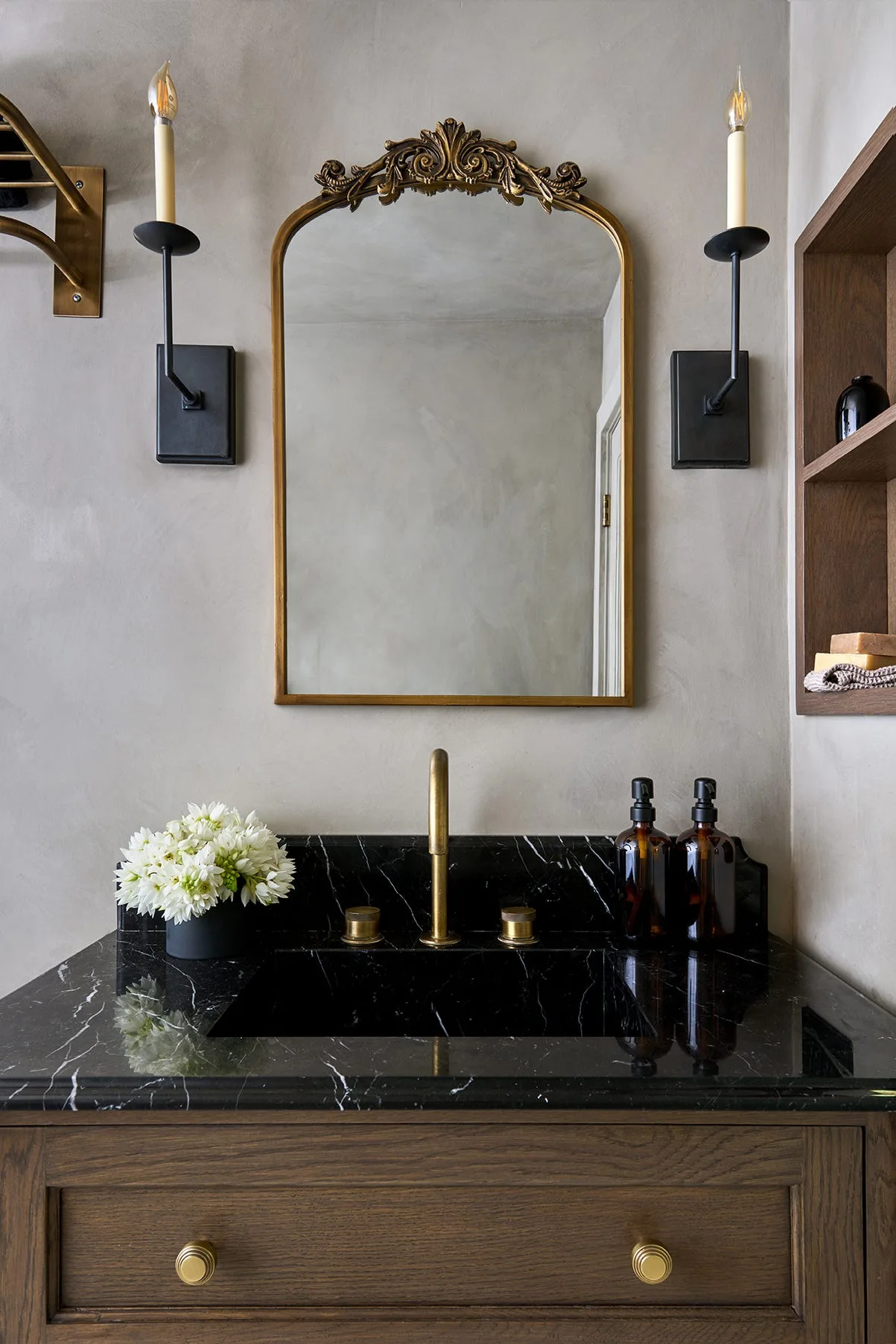 A bathroom vanity with a black marble countertop, a wood cabinet, a brass faucet, and an ornate gold mirror flanked by two candlestick sconces.