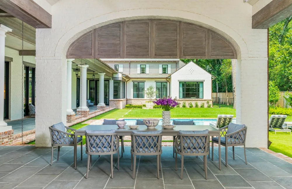A gray outdoor dining table and woven chairs on a stone patio under a brick archway overlooking a backyard pool.