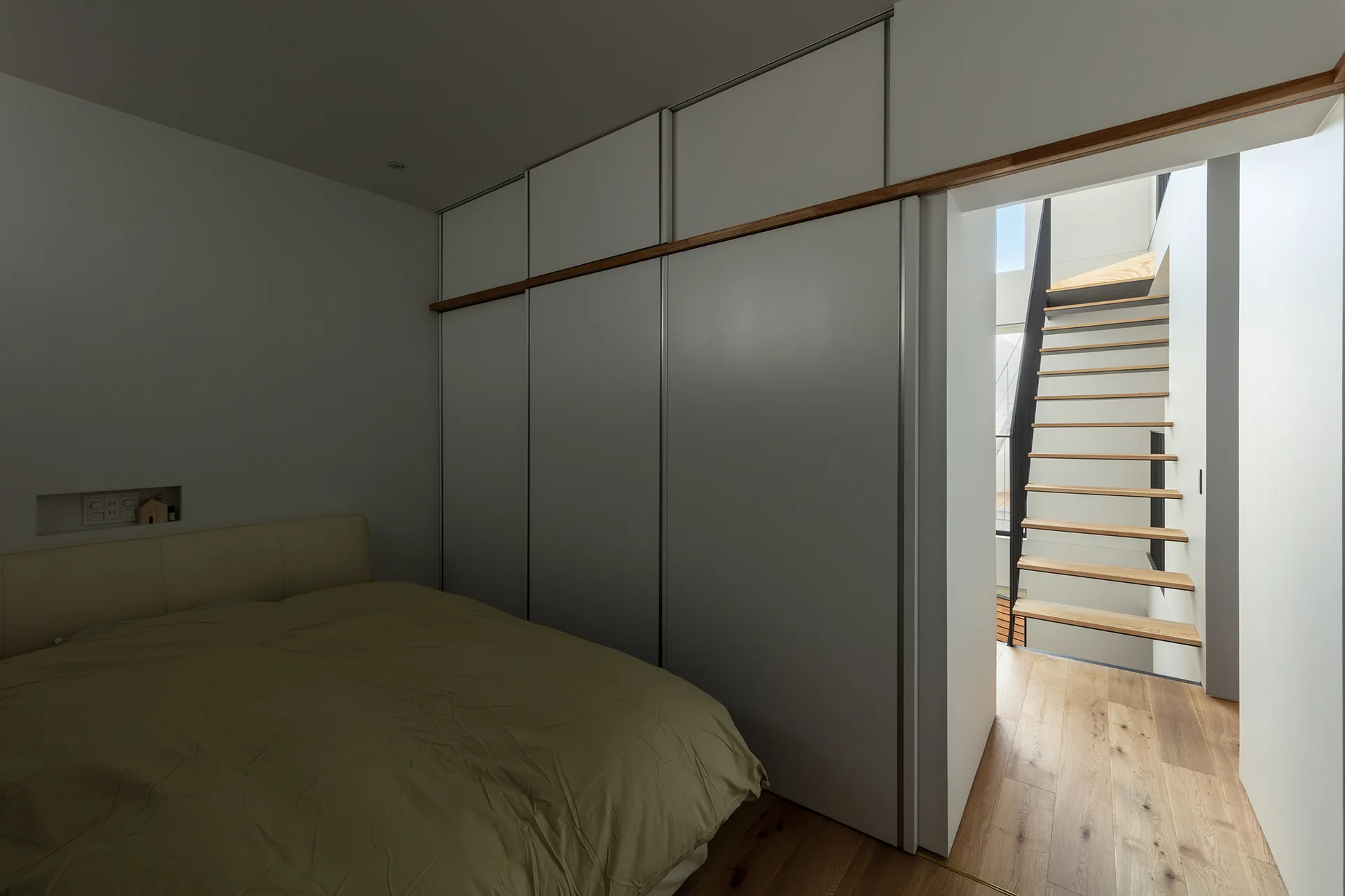 A minimalist bedroom features a low bed, large white floor-to-ceiling closet doors, and a doorway opening to an open-riser wood staircase.