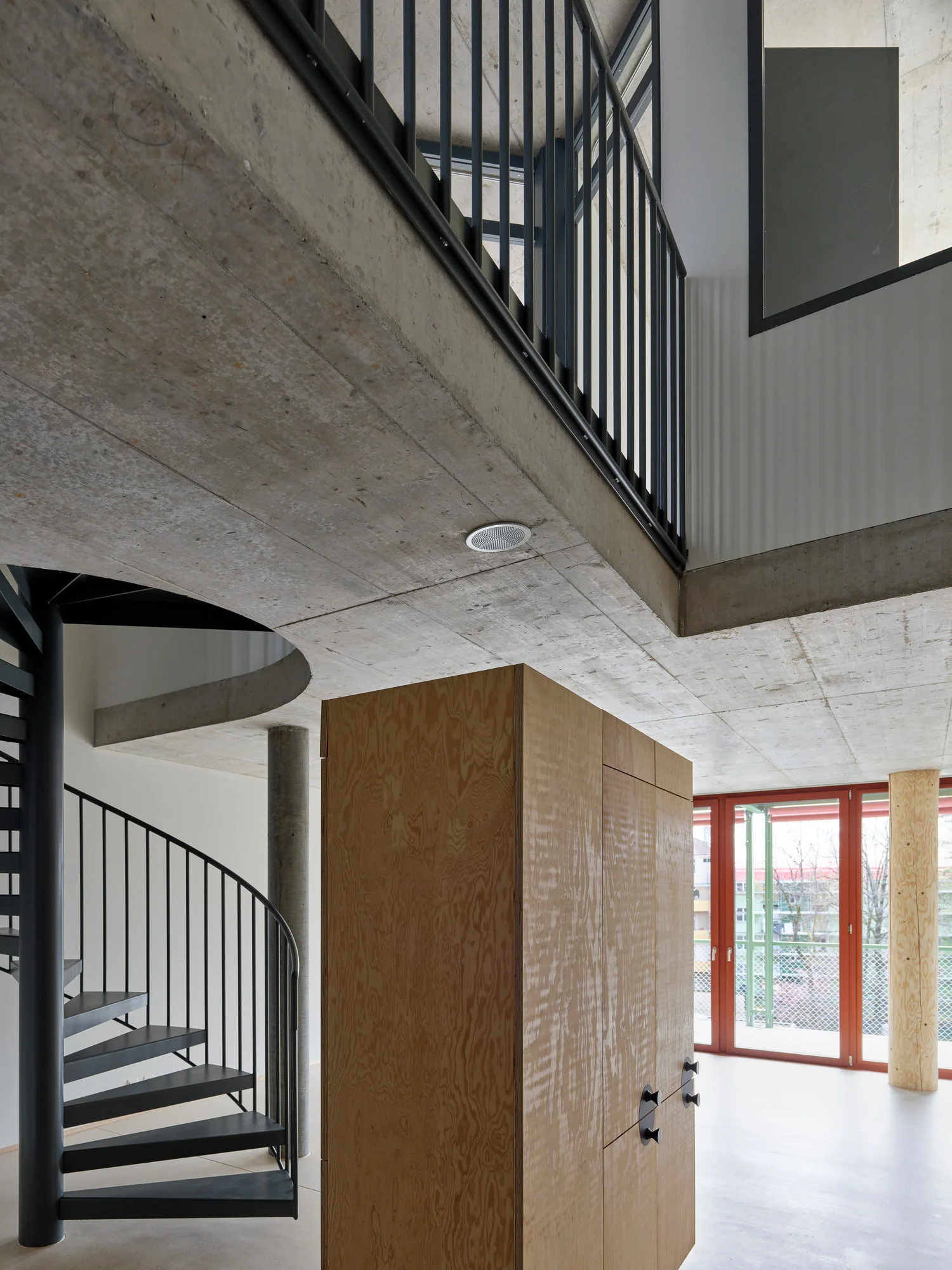 A modern industrial loft interior featuring a black metal spiral staircase, a large central plywood cabinet, and an exposed concrete ceiling with a mezzanine.