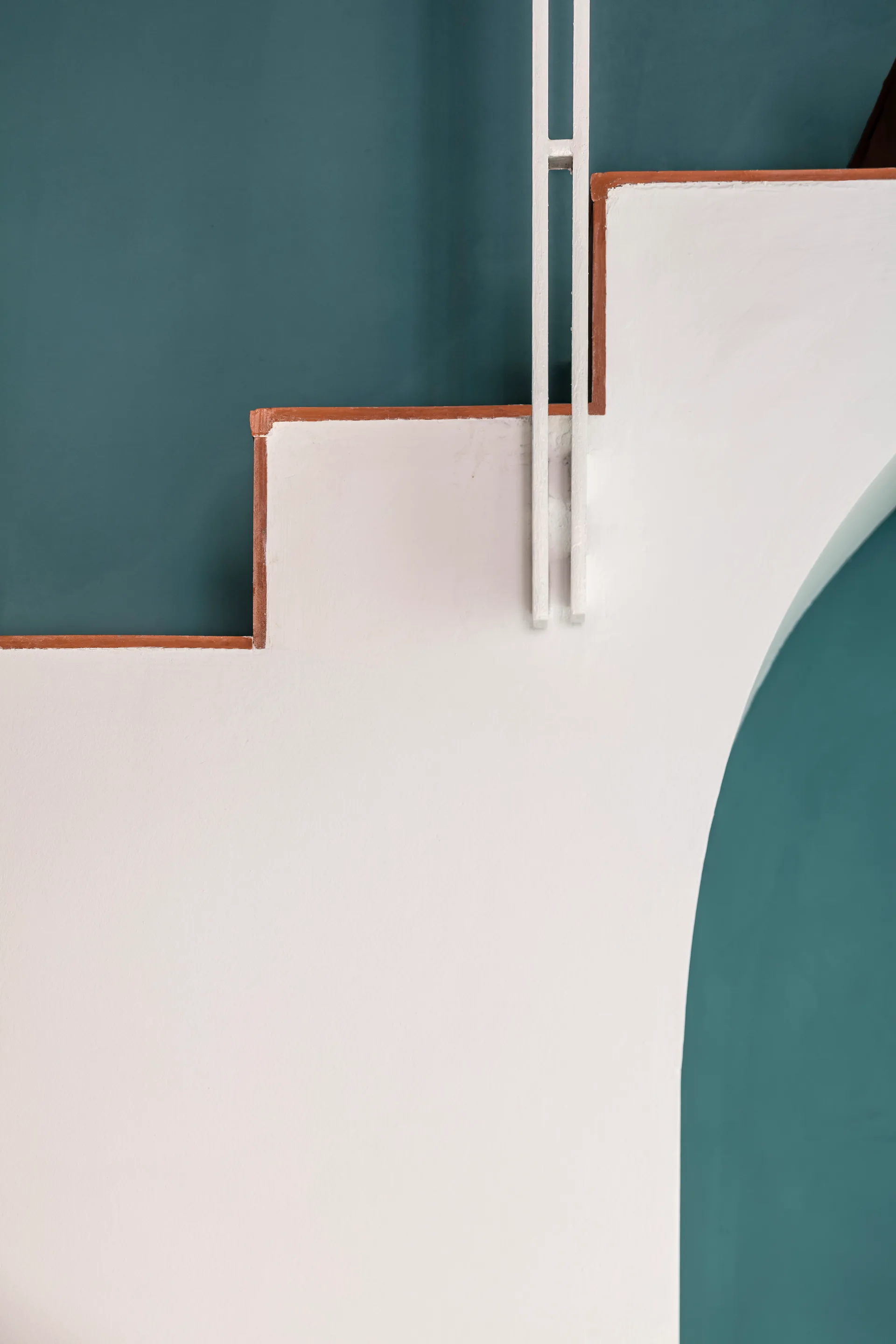 A minimalist white plaster staircase with a metal railing is shown against a matte teal wall with an archway.