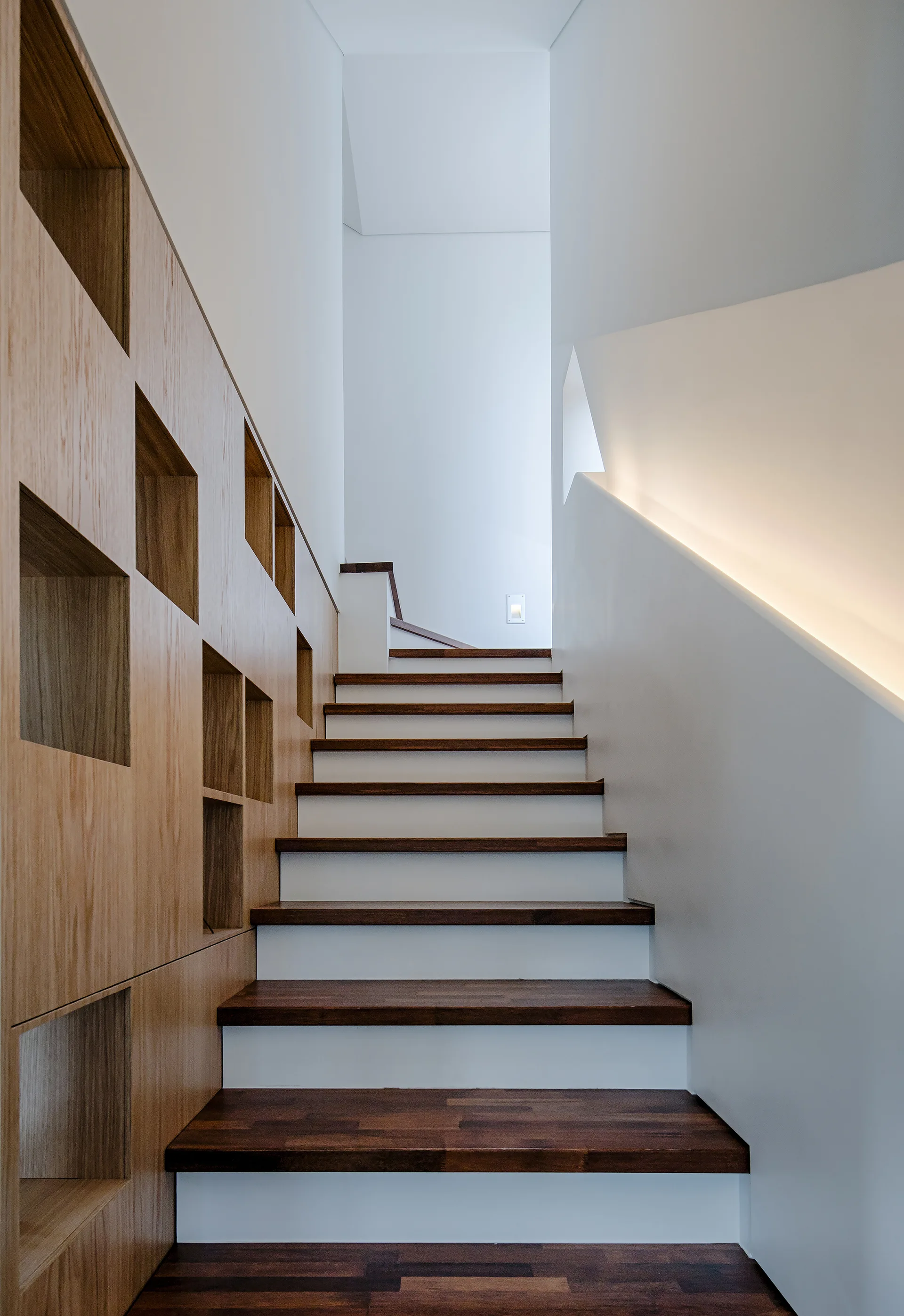 A minimalist staircase featuring dark wood treads and white risers next to a light wood paneled wall with recessed square display niches.