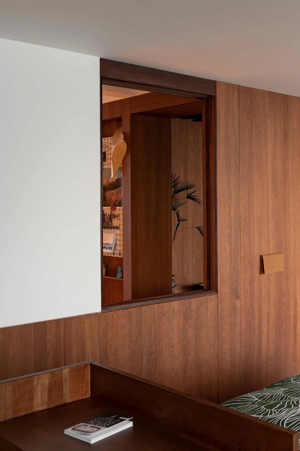 A vertical wood-paneled partition wall features a square indoor window cutout showing a glimpse of an adjacent room with a pegboard and plant.