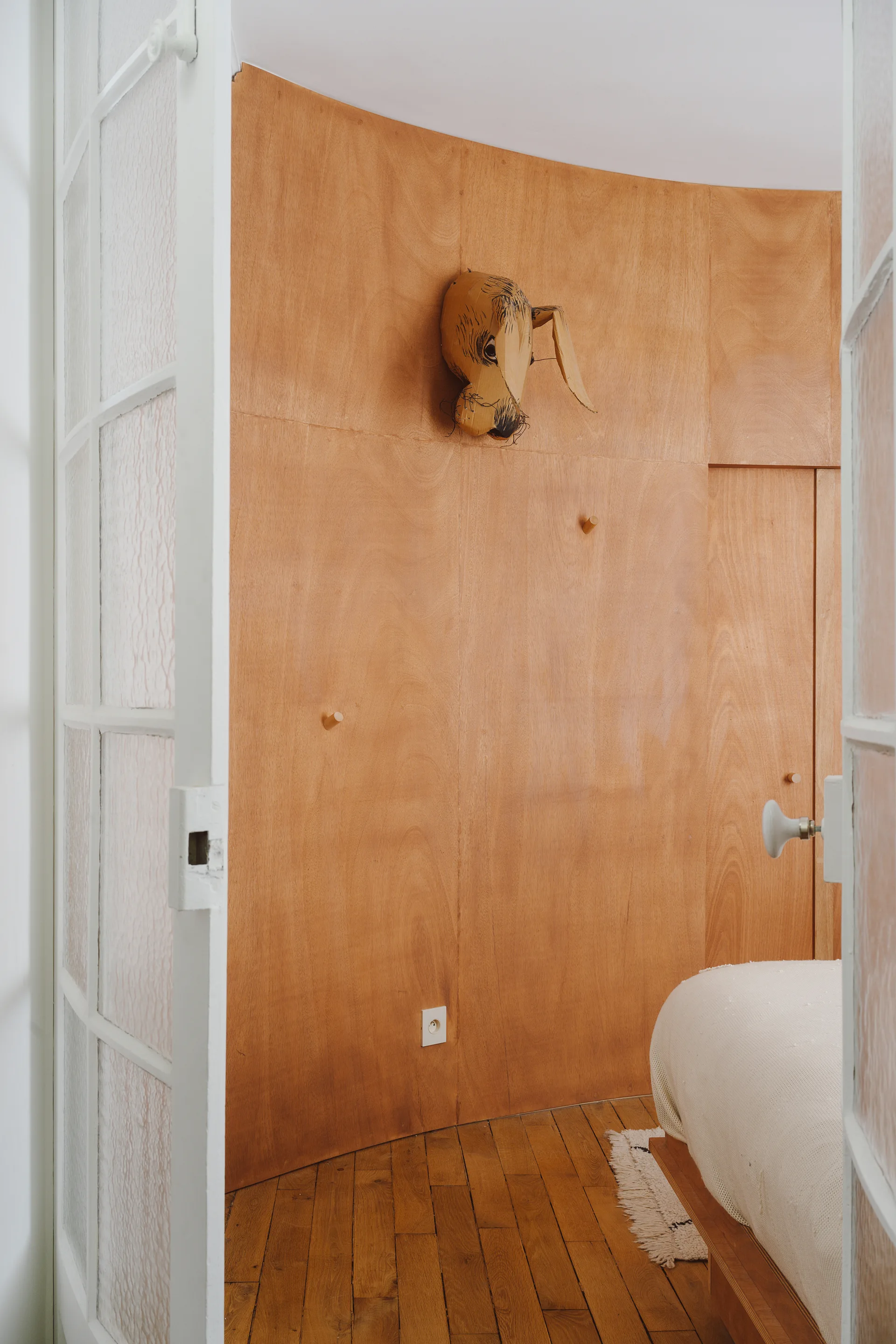 A view through a glass-paned door into a bedroom featuring curved plywood wall paneling, parquet wood floors, and a bed with white linens.