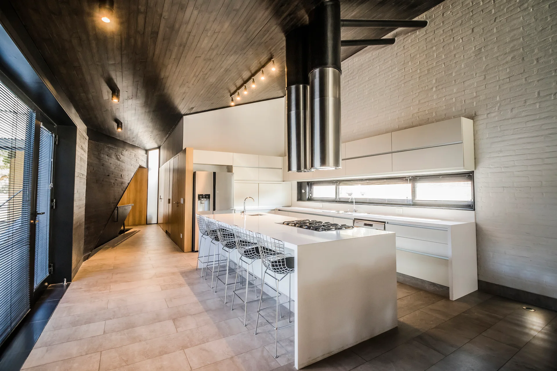 A modern kitchen featuring a long white island with wire stools, a dark wood plank ceiling, and dual stainless steel range hoods.