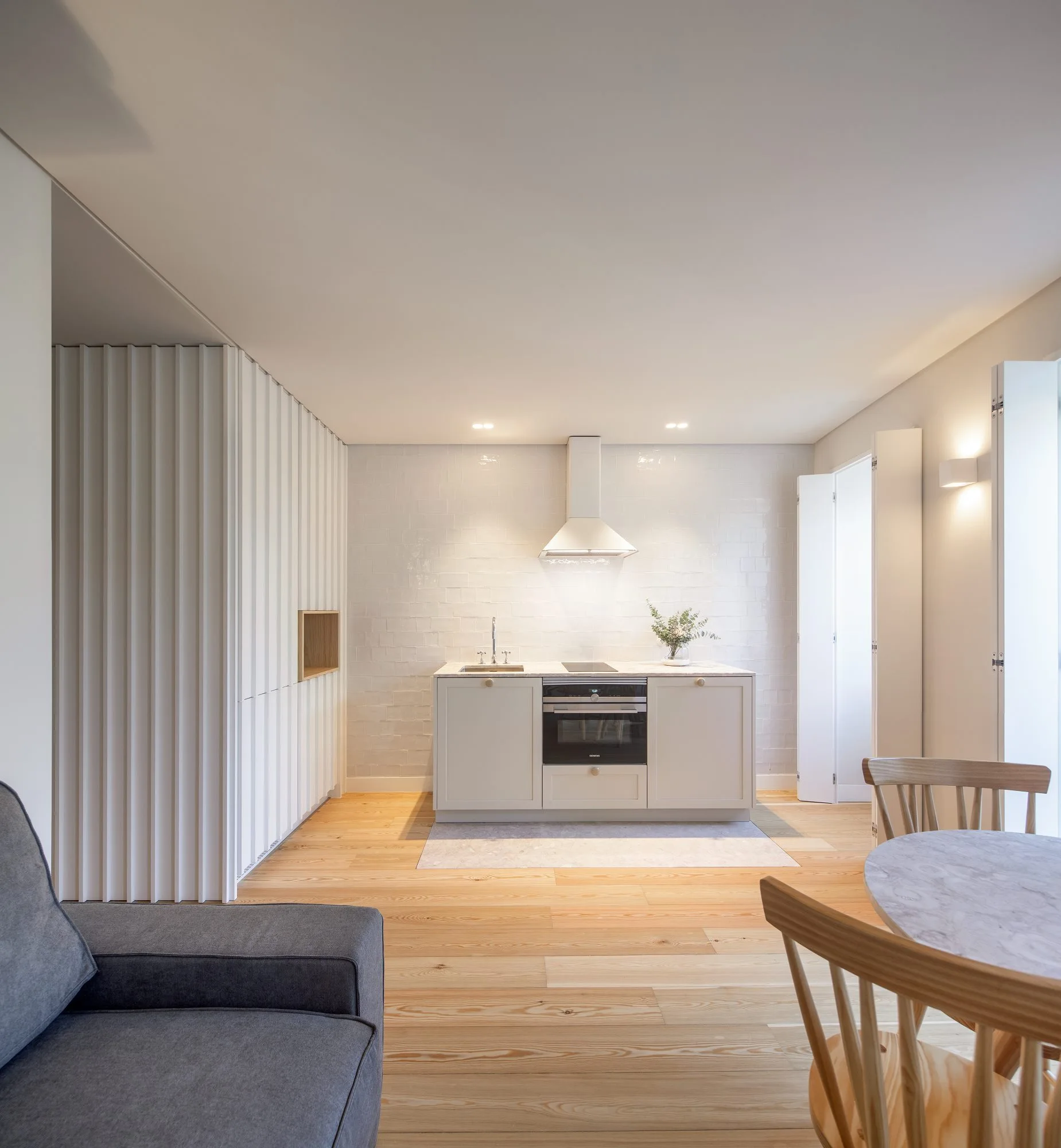 A minimalist open-plan kitchen features a compact island, a white glossy tile wall, and white vertical slatted paneling over light wood floors.