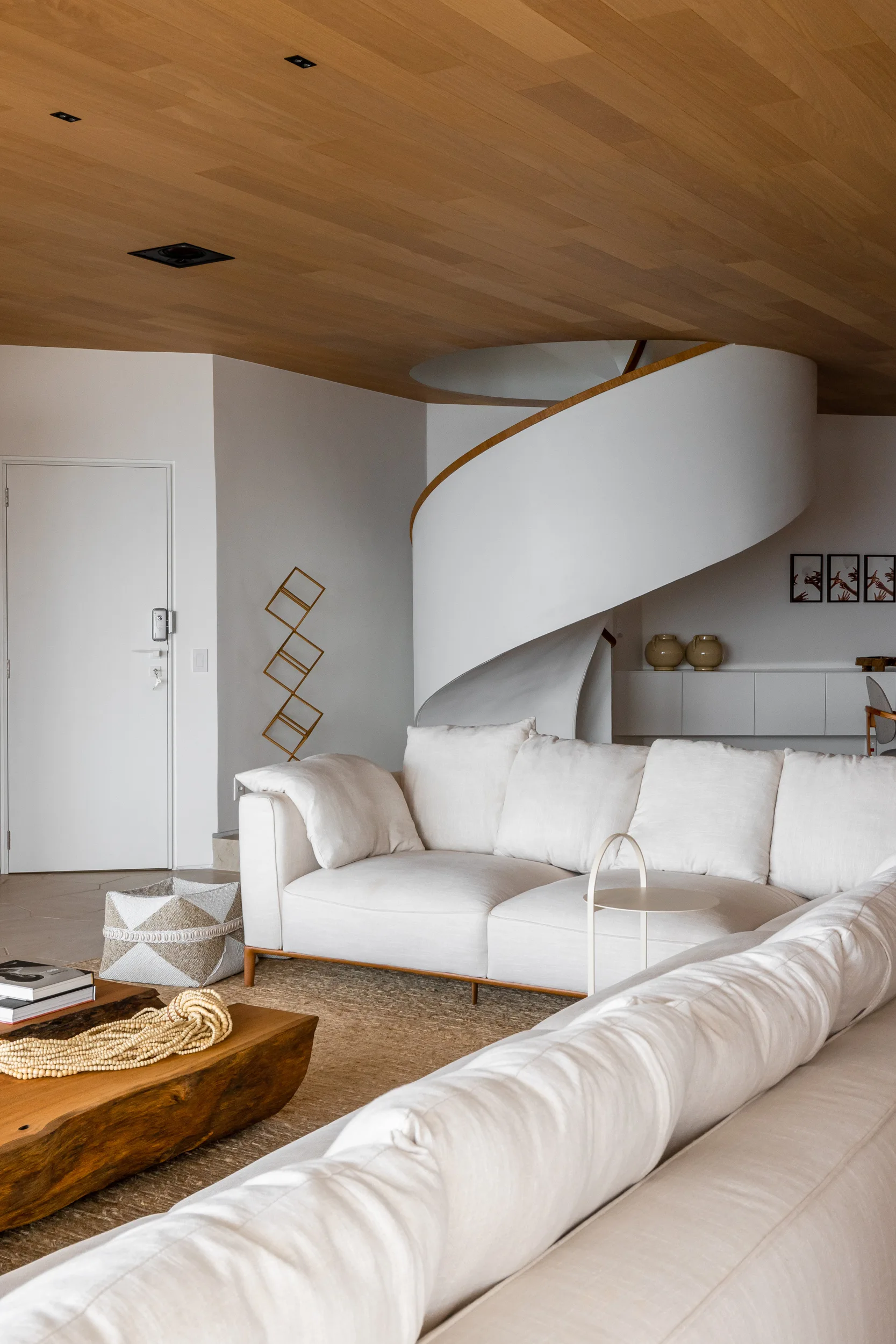 A minimalist living room features a cream sectional sofa, a live-edge wood coffee table, and a white curved staircase under a wood paneled ceiling.