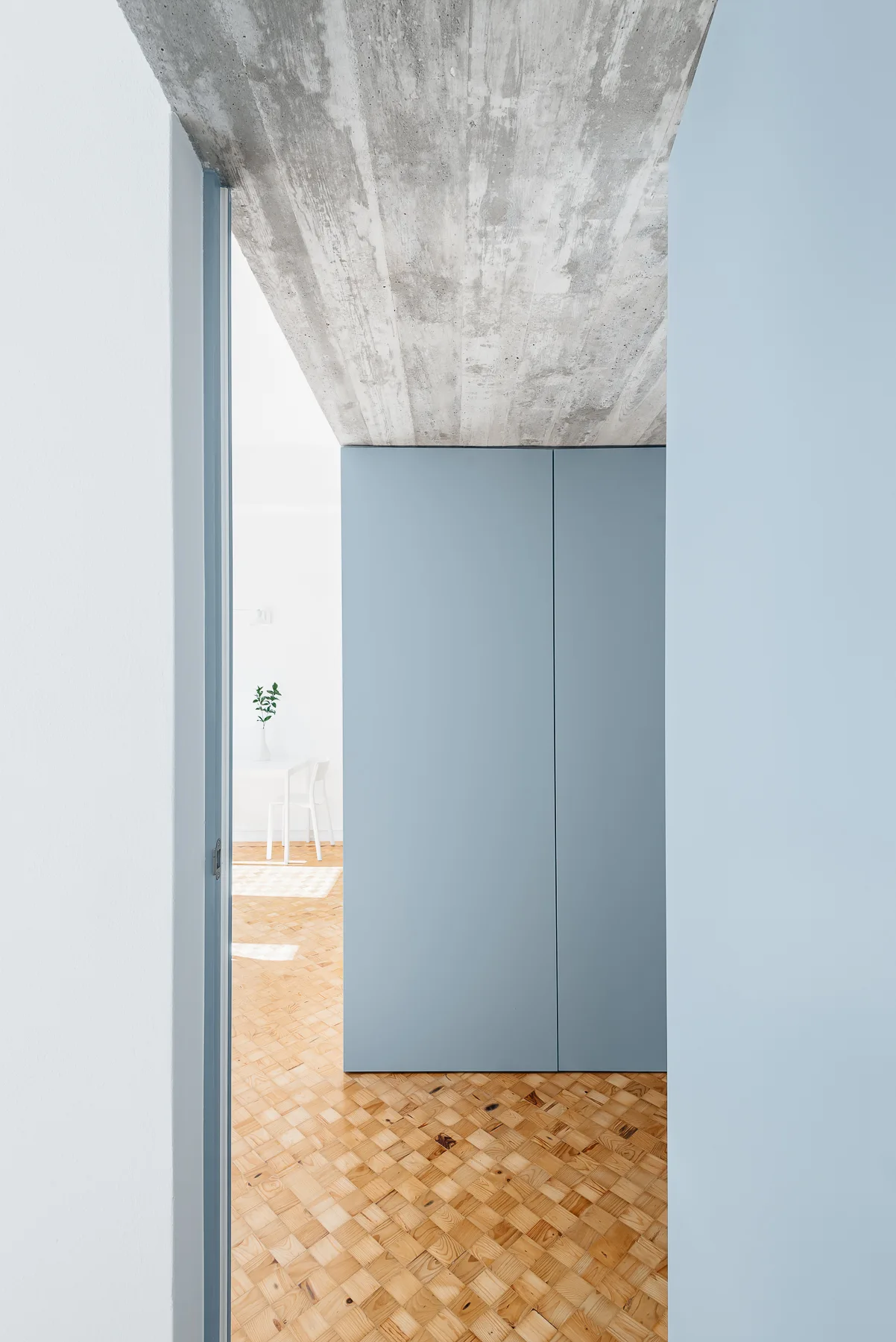 A minimalist hallway features matte blue built-in cabinets, an exposed concrete ceiling, and warm wood parquet flooring leading into a white room.