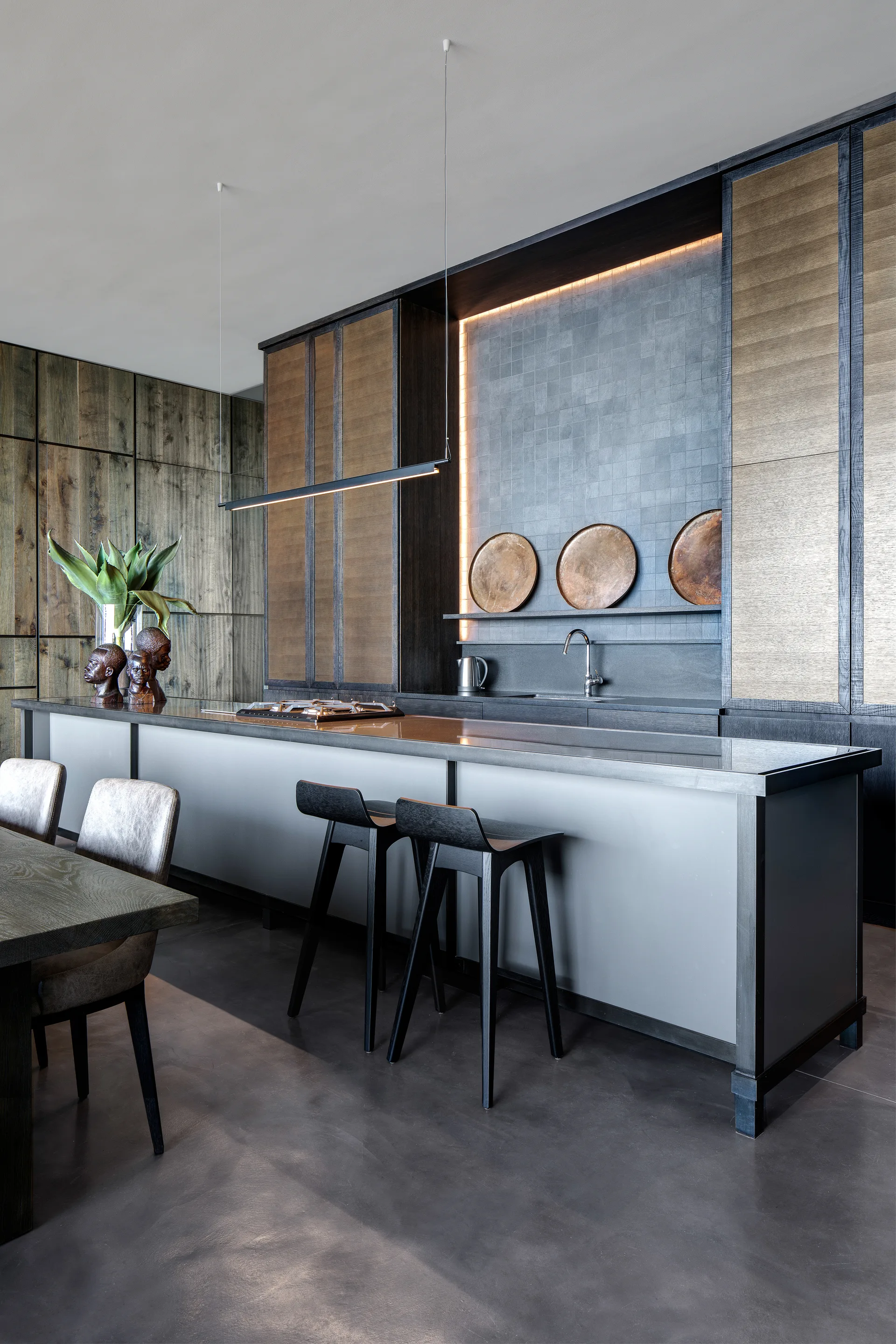 A contemporary kitchen with a long charcoal island, black bar stools, dark wood-grained cabinets, and a tiled backsplash area with three copper plates.