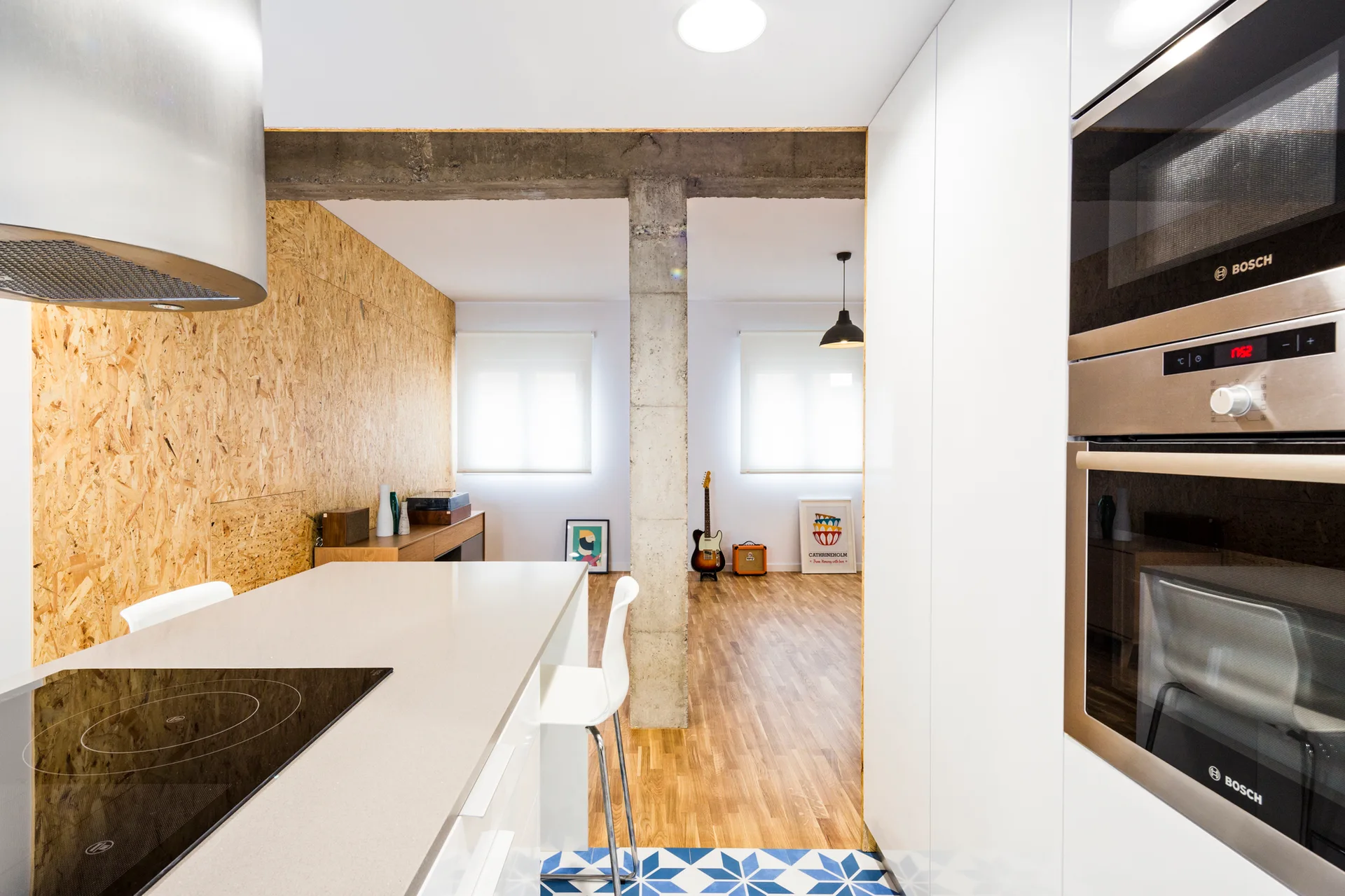 A modern white kitchen with a concrete column, OSB paneled wall, and blue geometric tile flooring transitioning to a wood-floored living area.