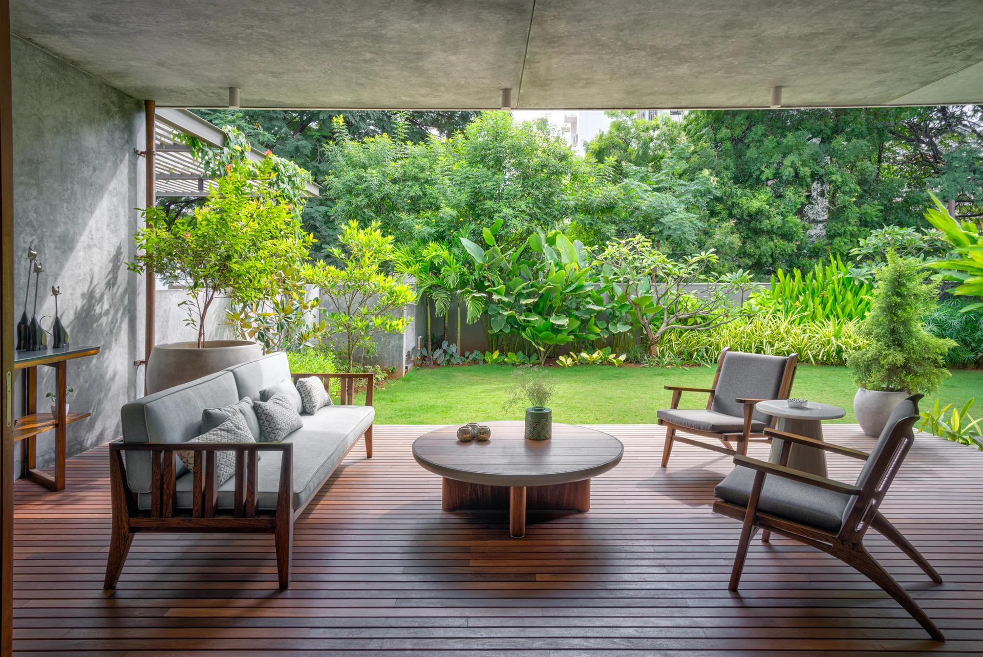 A covered concrete patio features wood plank decking, a gray cushioned sofa, two matching armchairs, and an open view of a lush green backyard garden.