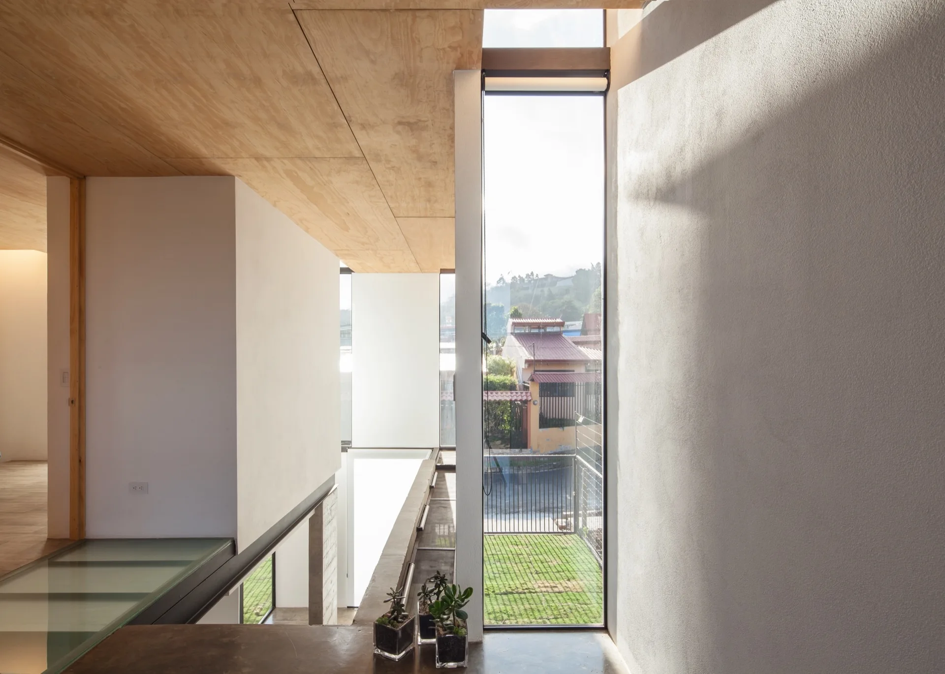 A minimalist landing features a light plywood ceiling, white walls, a glass floor panel, and a tall vertical window overlooking a residential street.