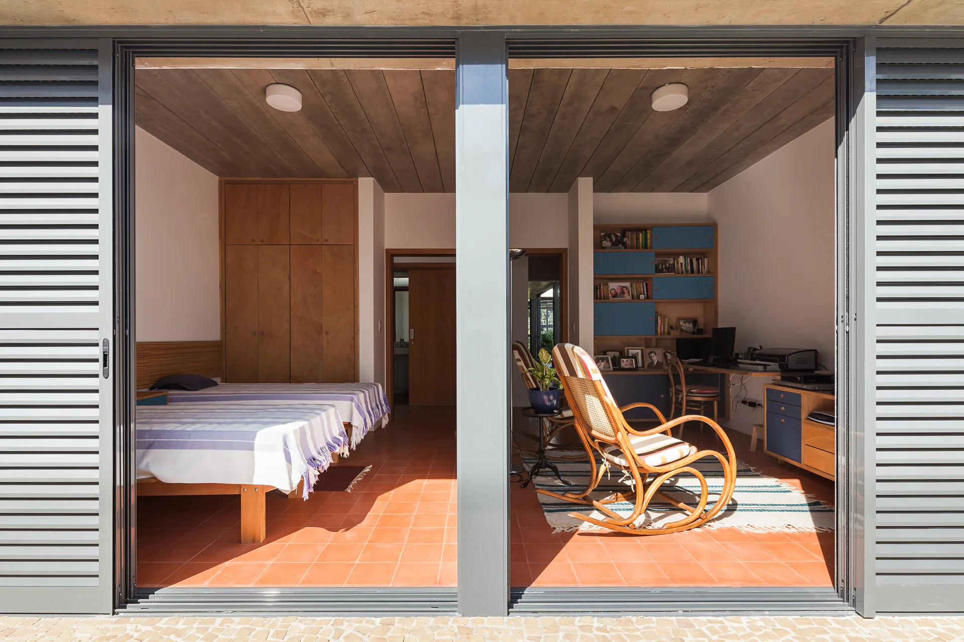 A view through sliding glass doors into a bedroom and study with terracotta tile floors, wooden beds, a rattan rocking chair, and built-in shelving.