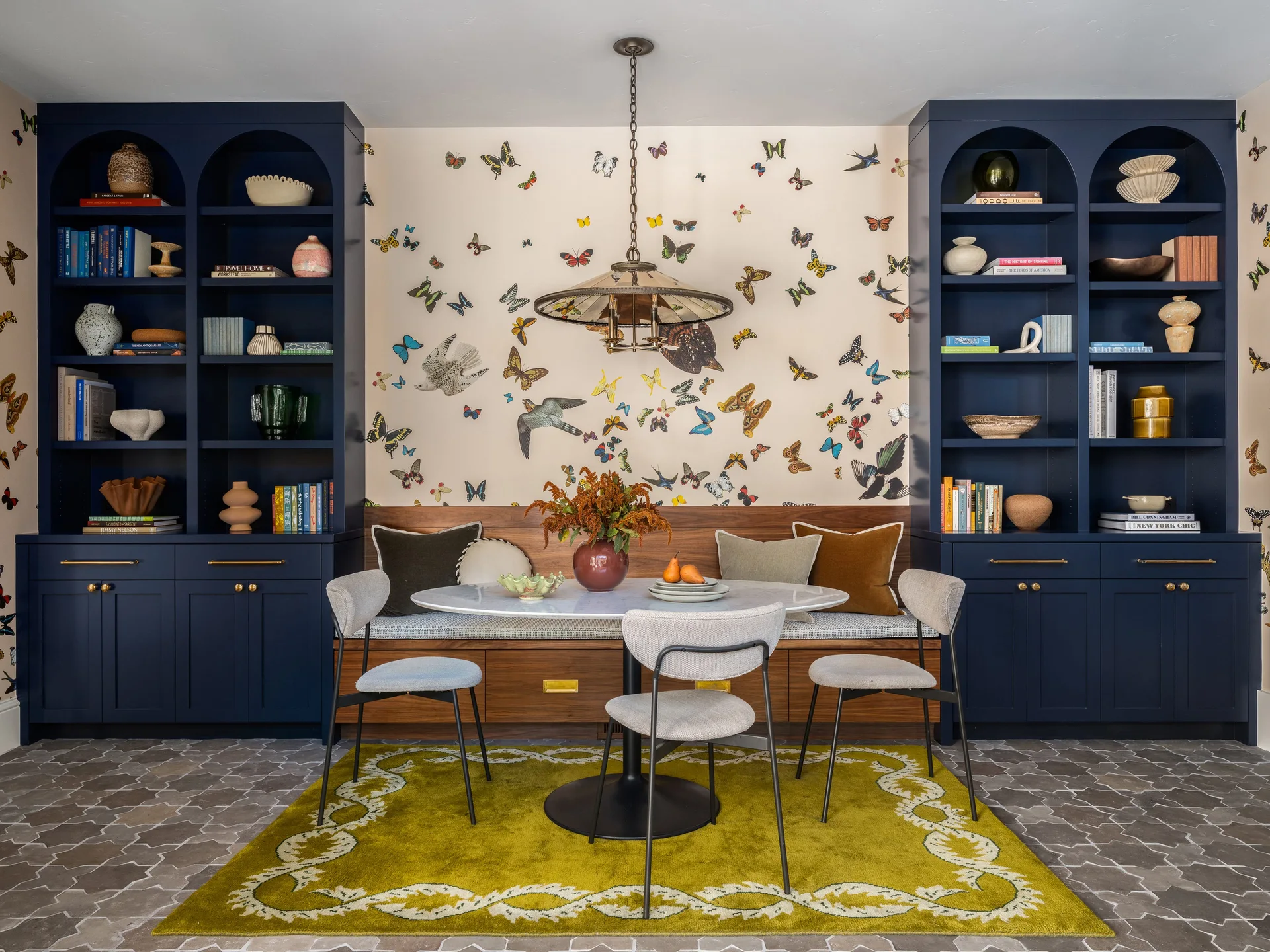 A dining area features symmetrical navy blue arched bookcases flanking a wood banquette niche with butterfly wallpaper, a marble table, and a green rug.