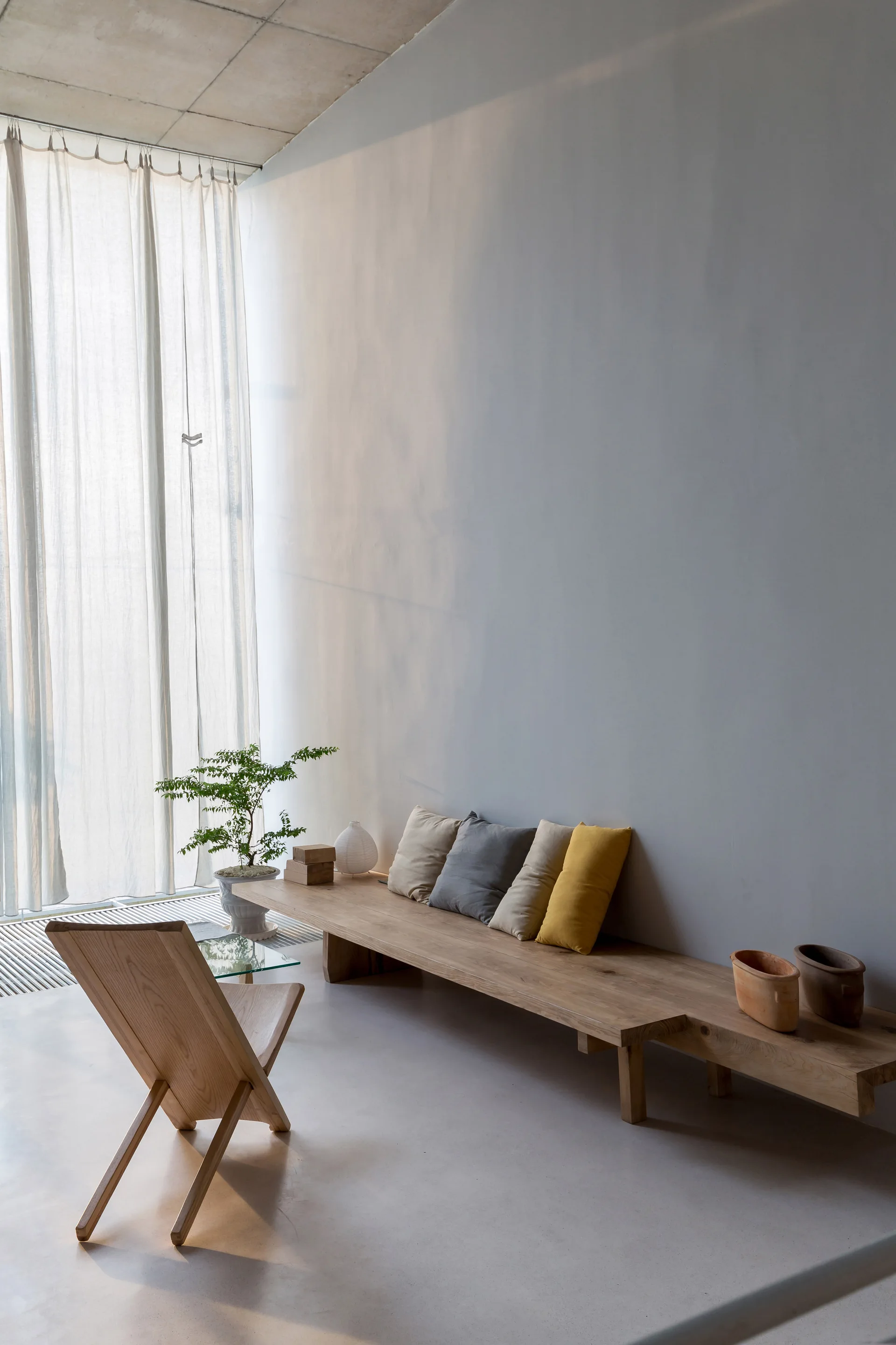 A minimalist living room features a long wooden bench with throw pillows, a low-slung wooden chair, and sheer white curtains filtering natural light.