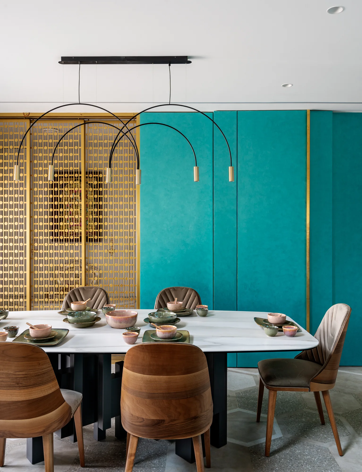 A contemporary dining room features a white marble table, wood-backed chairs, a sculptural black pendant light, and a teal and gold paneled backdrop.