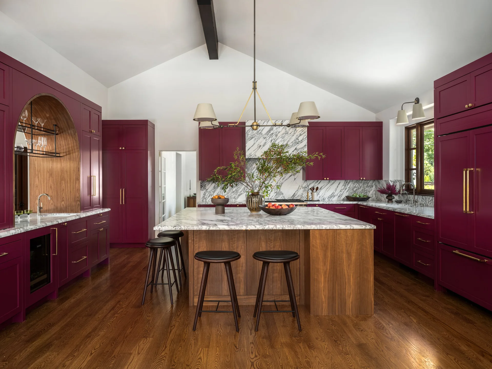 A kitchen featuring deep burgundy cabinets, a marble-topped wood island with bar stools, an arched wood niche, and a vaulted ceiling.