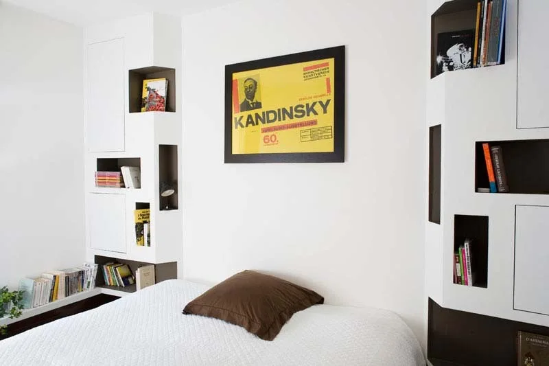 A bed with a textured white coverlet is flanked by custom white built-in cabinets featuring dark recessed niches and book storage.