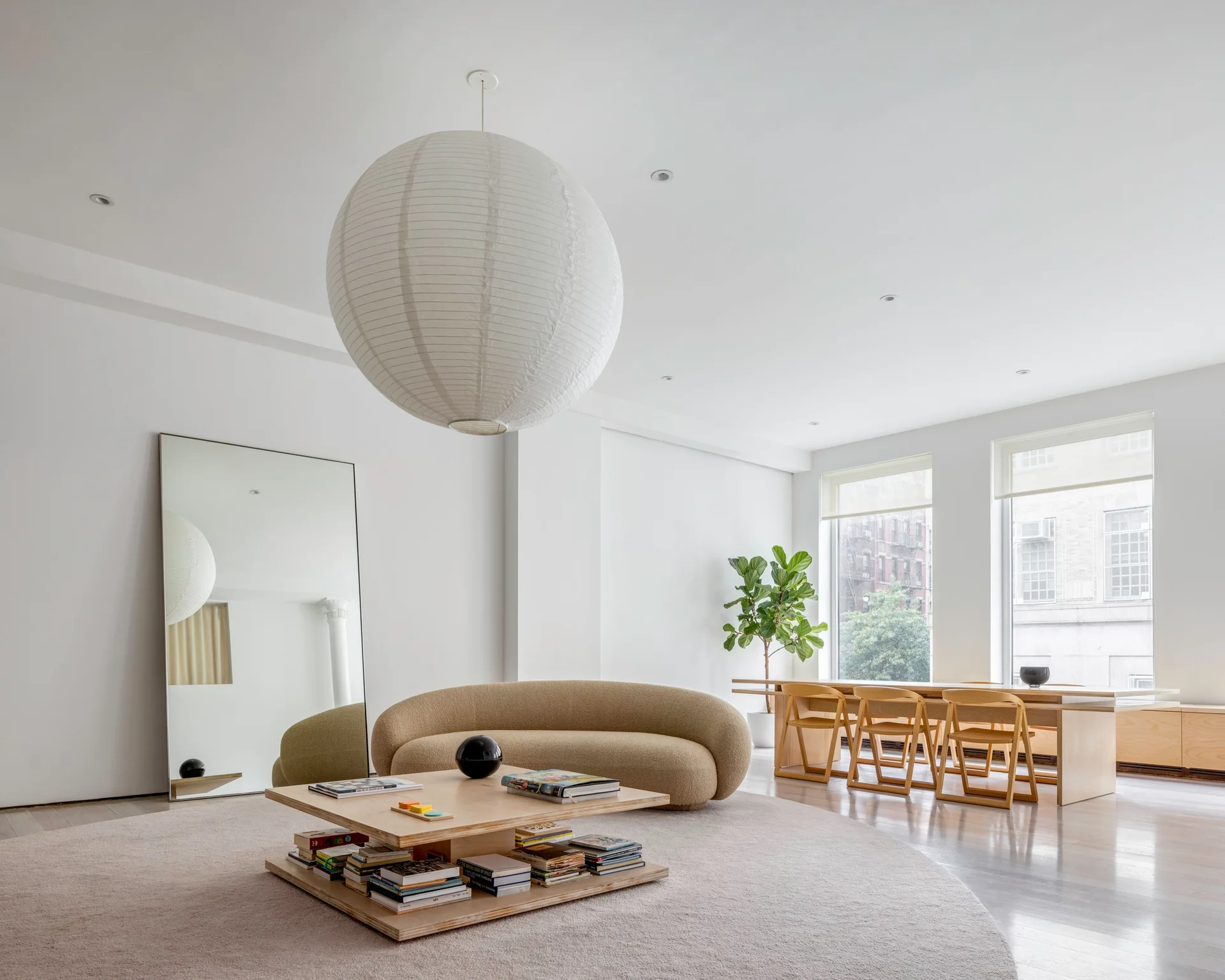 A minimalist living space featuring a curved beige sofa, a tiered wooden coffee table, and an oversized round paper pendant light.