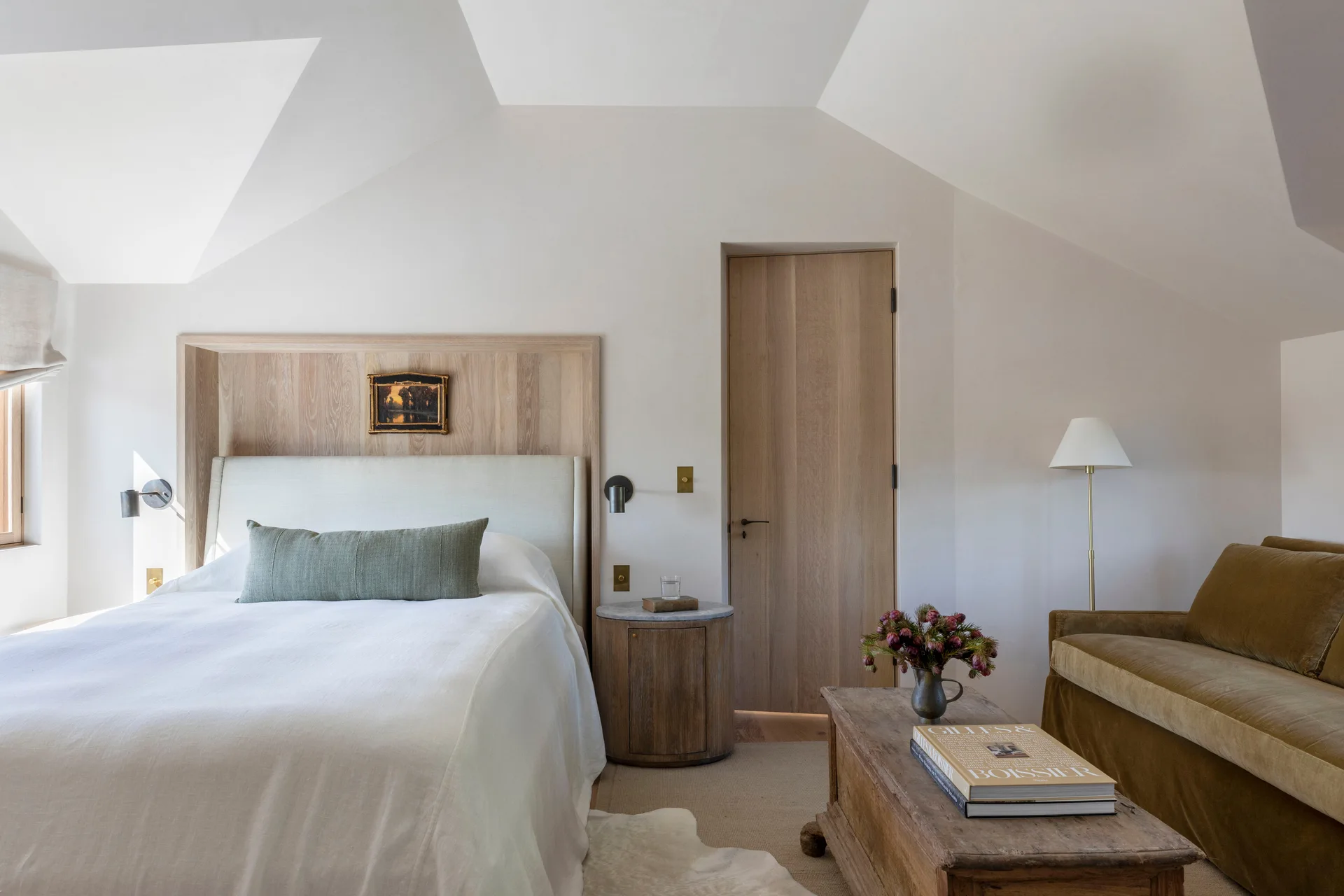 A neutral bedroom features a bed with a light wood panel headboard, a matching wood flush door, a velvet sofa, and an angled sloped ceiling.