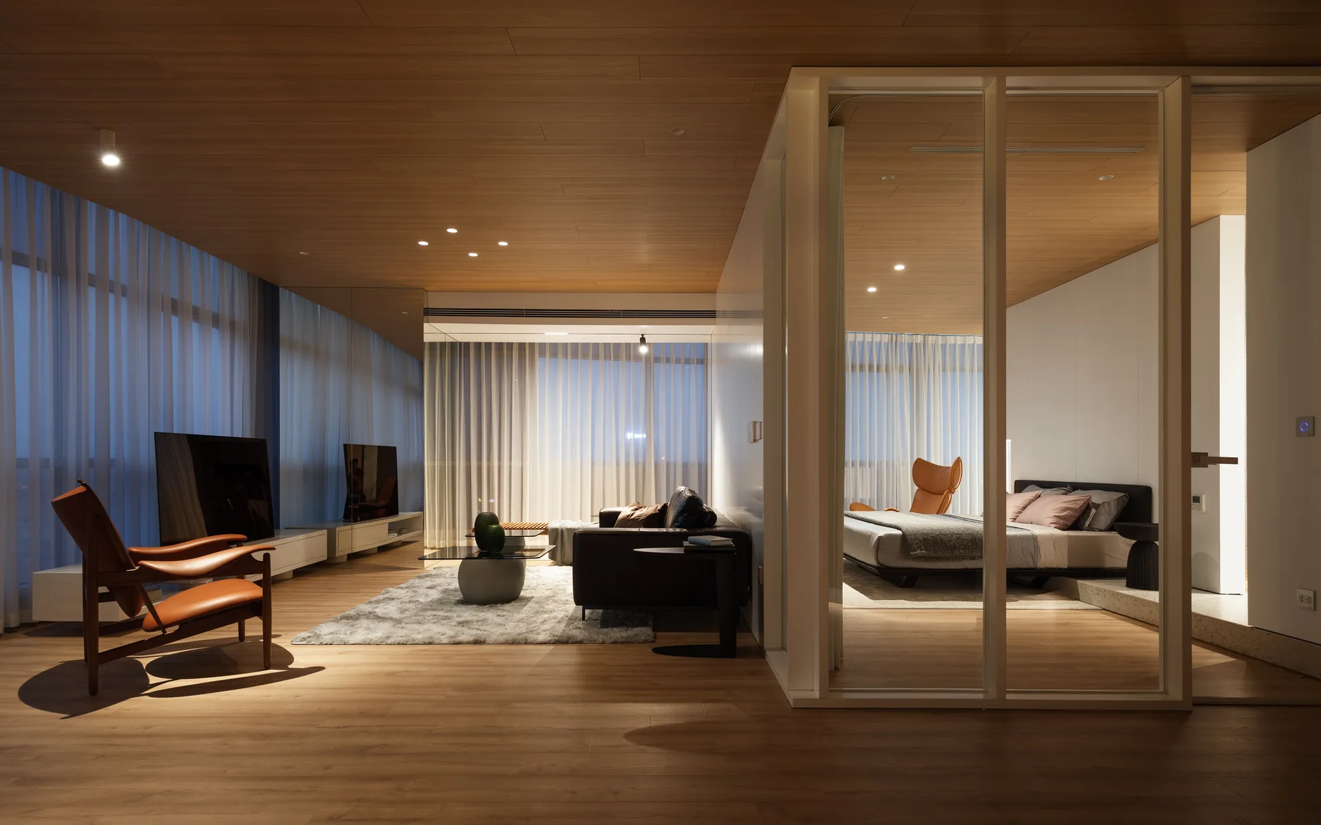 An open-concept living area and bedroom divided by a white-framed glass partition, featuring wood plank ceilings, a leather sofa, and sheer curtains.