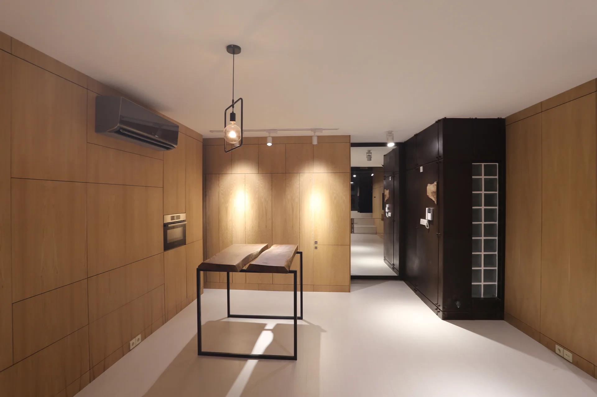 A minimalist kitchen features wood paneled walls, an integrated oven, a slim live edge wood island table, and a dark metal cabinet with glass blocks.