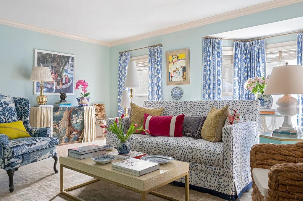 A light blue living room features a patterned sofa with red and gold pillows, patterned blue curtains, and a gold-framed coffee table on a neutral carpet.