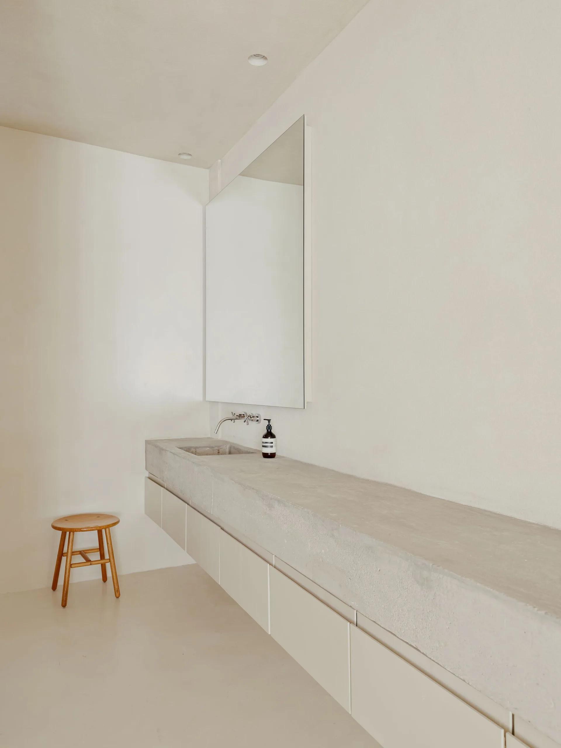A minimalist bathroom features a long wall-mounted concrete vanity with an integrated sink, a large frameless mirror, and a small wooden stool.