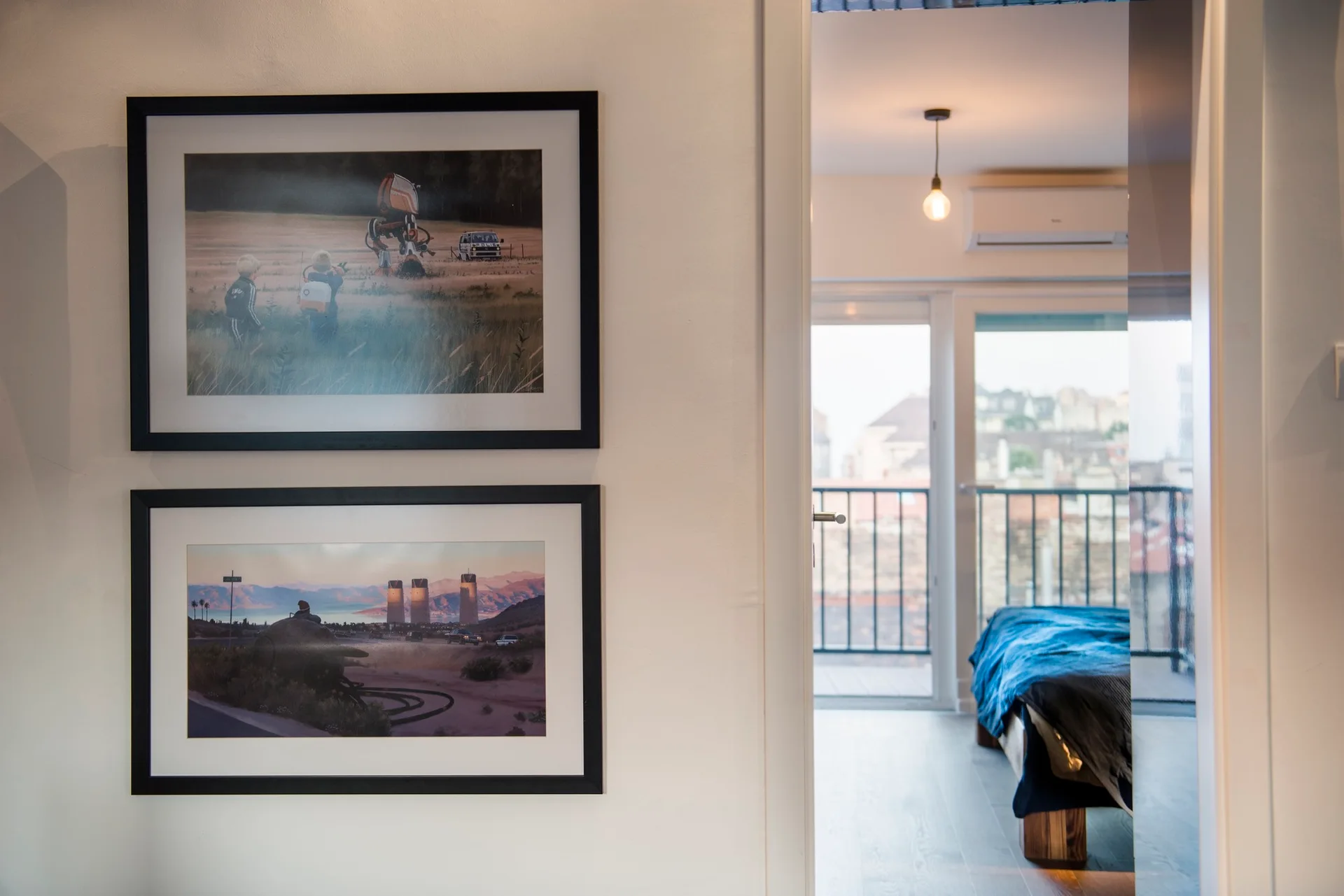 Two vertically stacked framed prints hang on a white wall next to an open doorway revealing a minimalist bedroom with a blue bed cover and balcony.