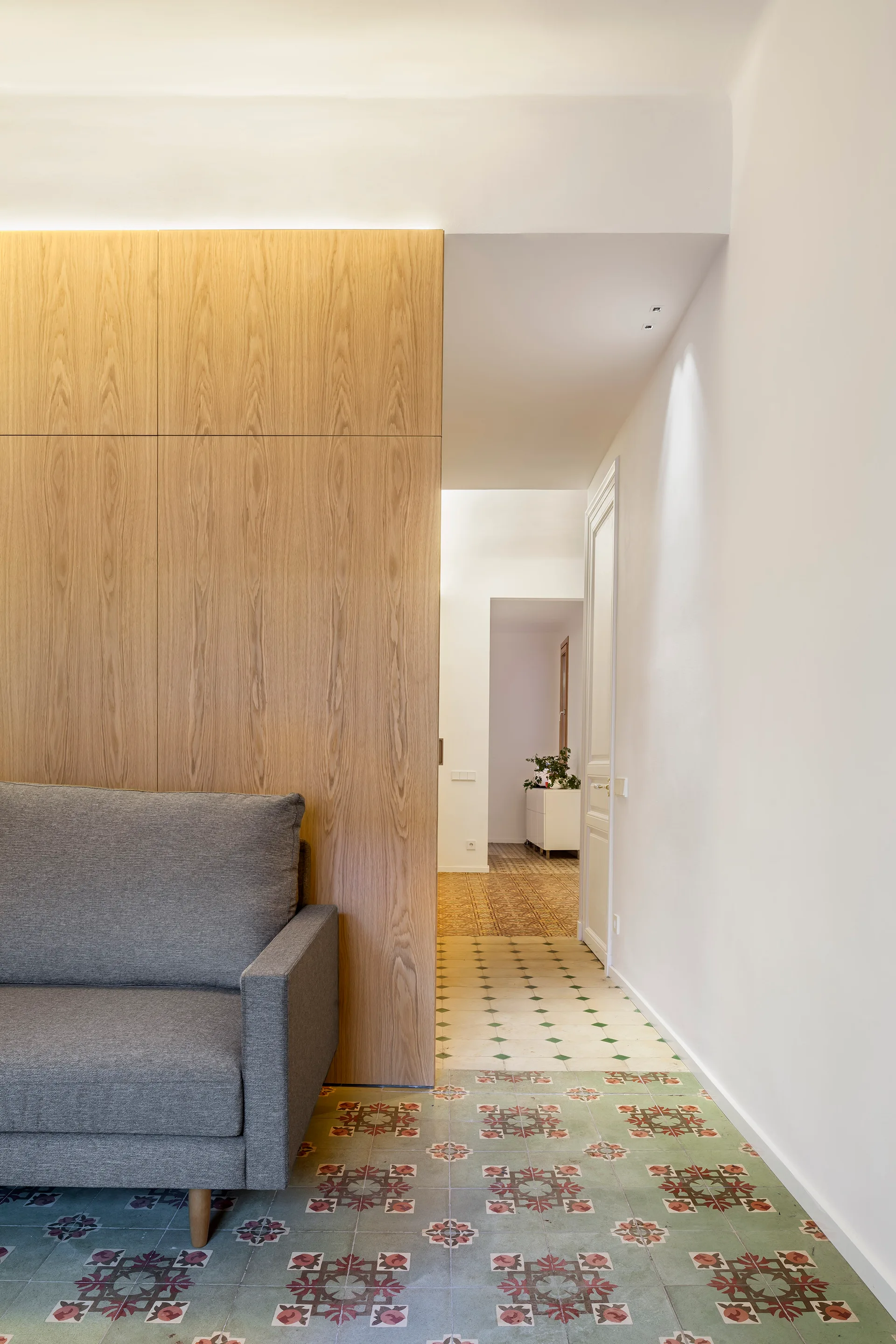 A grey fabric sofa stands next to a vertical wood panel divider on geometric patterned floor tiles leading down a white hallway.