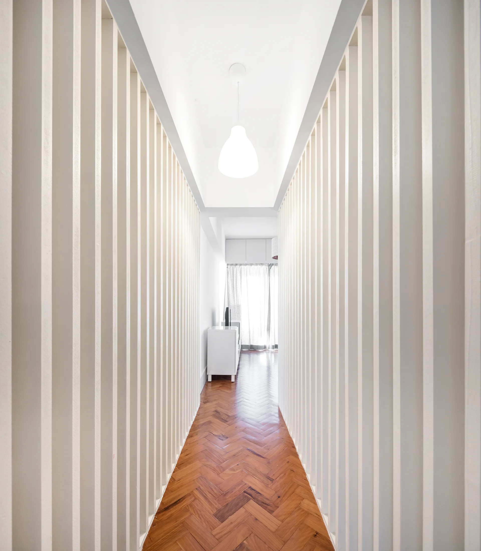 A narrow white hallway features vertical slatted wall panels, a herringbone wood floor, and a single pendant light hanging from a recessed ceiling.