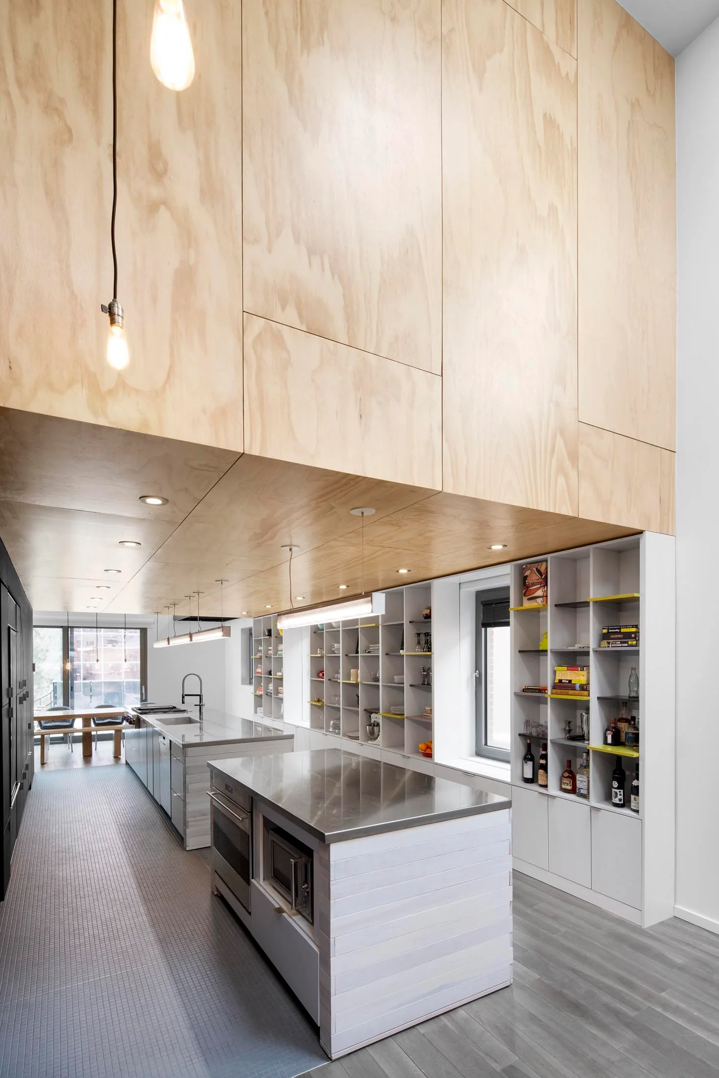 A modern kitchen featuring a warm plywood soffit overhead, stainless steel island countertops, and a full wall of white open shelving.