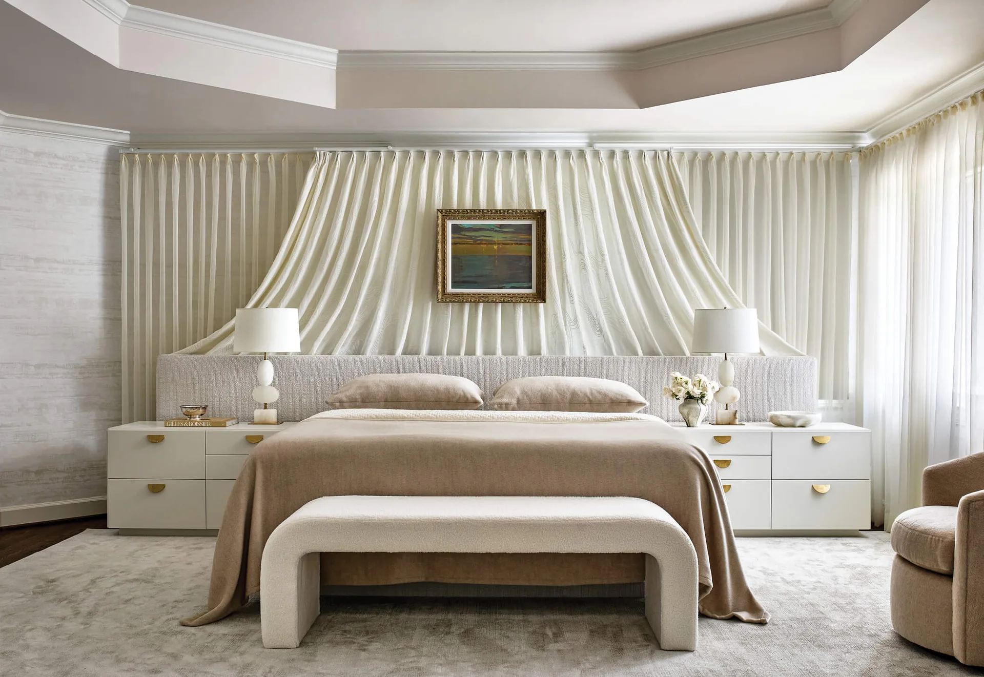 A cream bedroom featuring a bed with a low upholstered headboard set against a wall of draped white curtains, flanked by white nightstands.